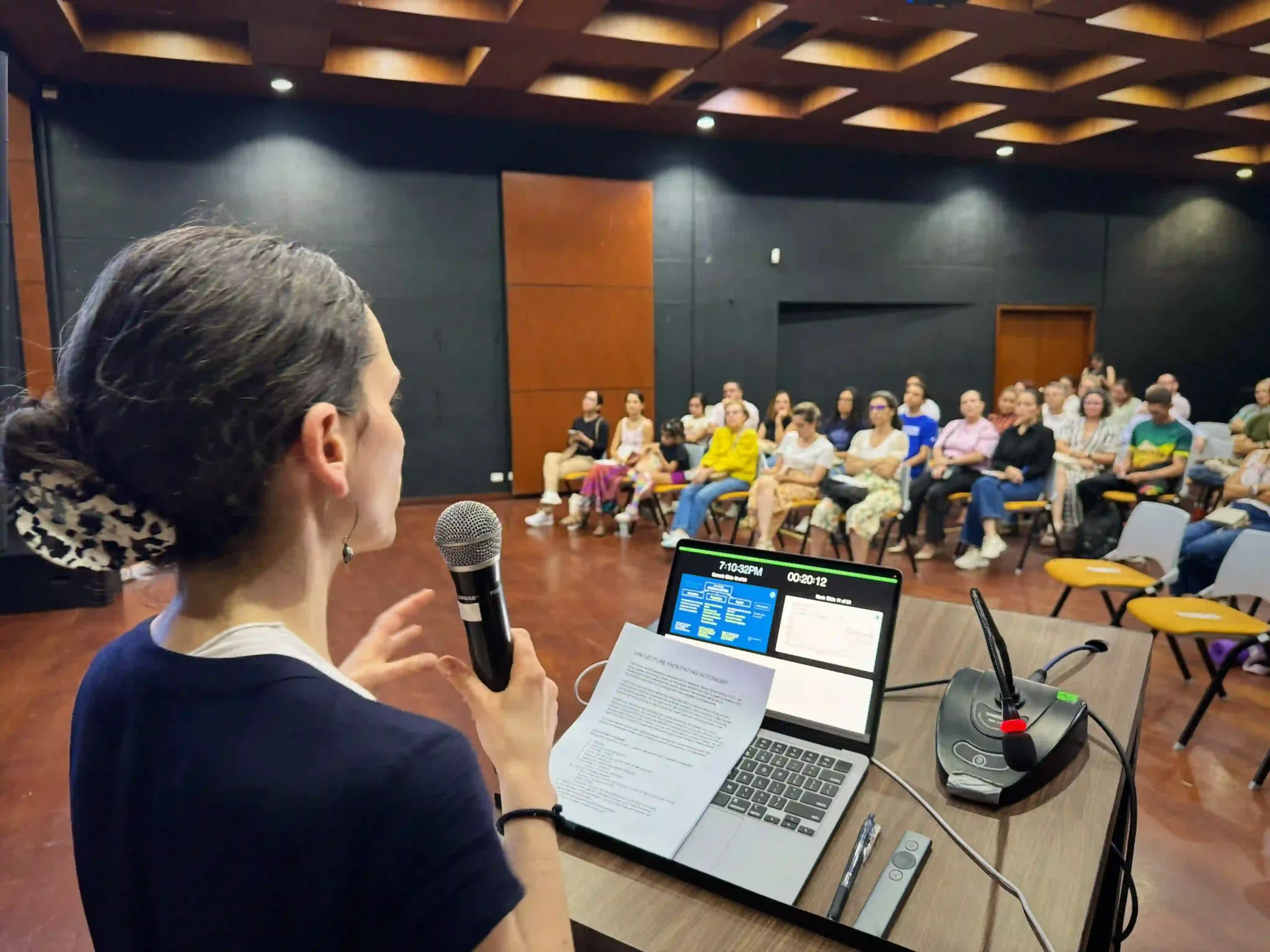 A female presenter in front of a lectern holds a microphone and speaks in front of an audience of people seating in chairs in a room with dark walls