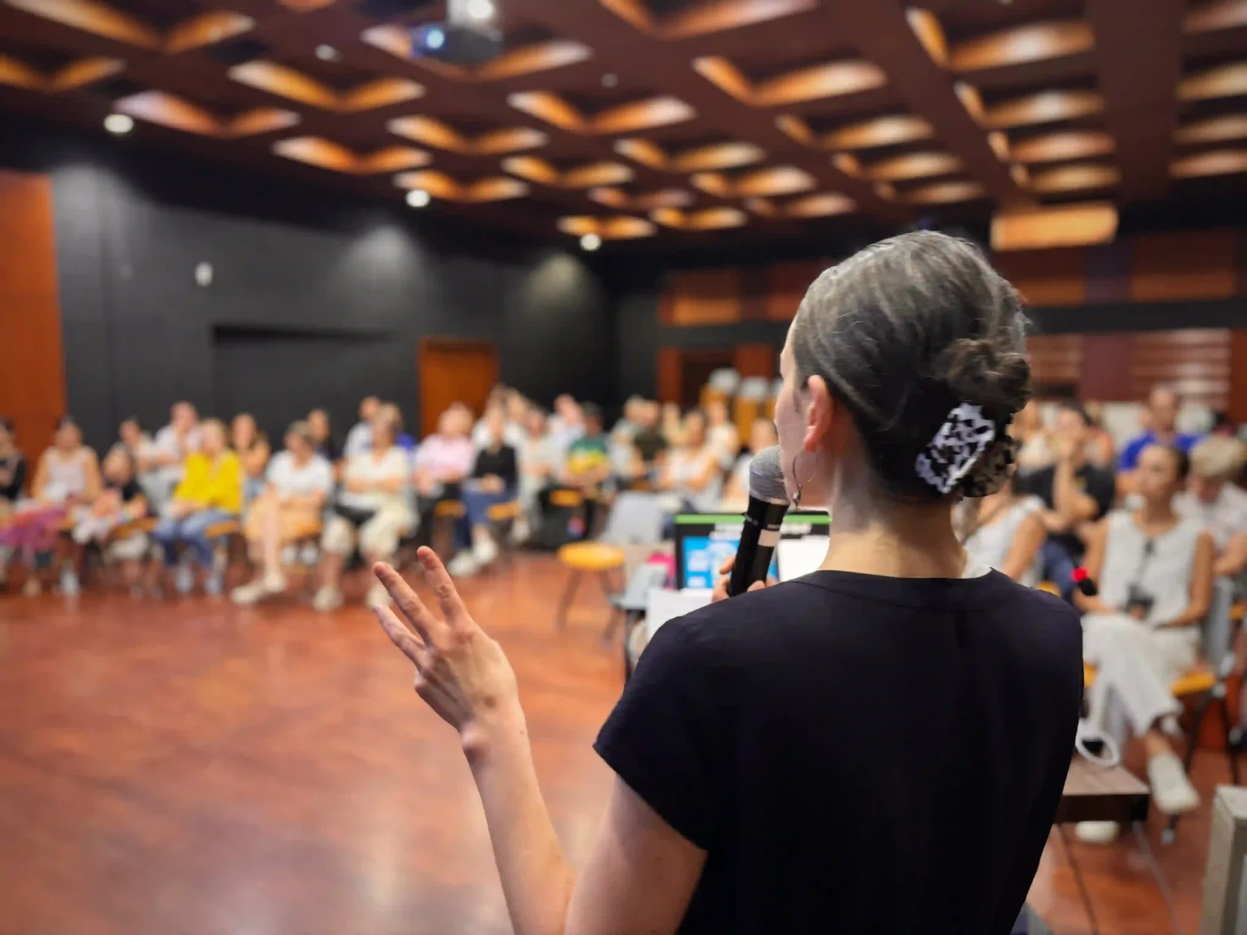 View from behind: a female presenter holds a microphone and shows number 3 with her fingers, speaking about the 3 types of body awareness, in front of an audience of people seated in chairs in a room with dark walls