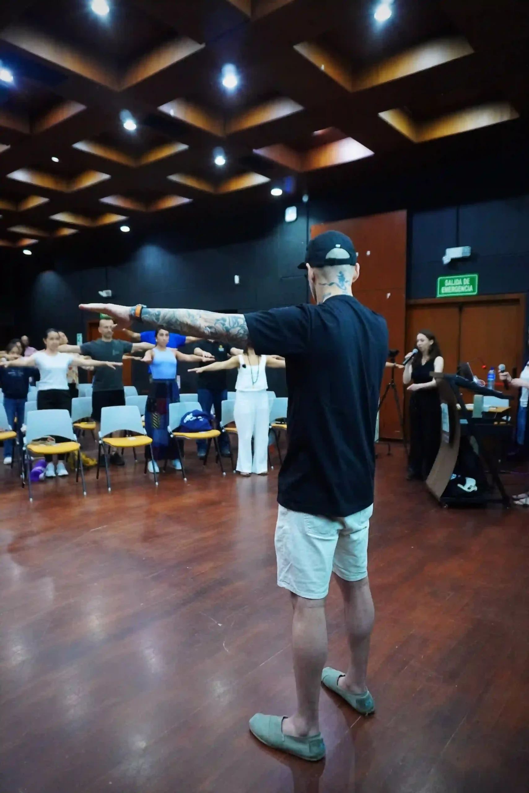 A male instructor with tattoed arms wearing a black T-shirt and a black cap stands with his arms out and instructing the movement to a group of people who stood up from their chairs in a conference room