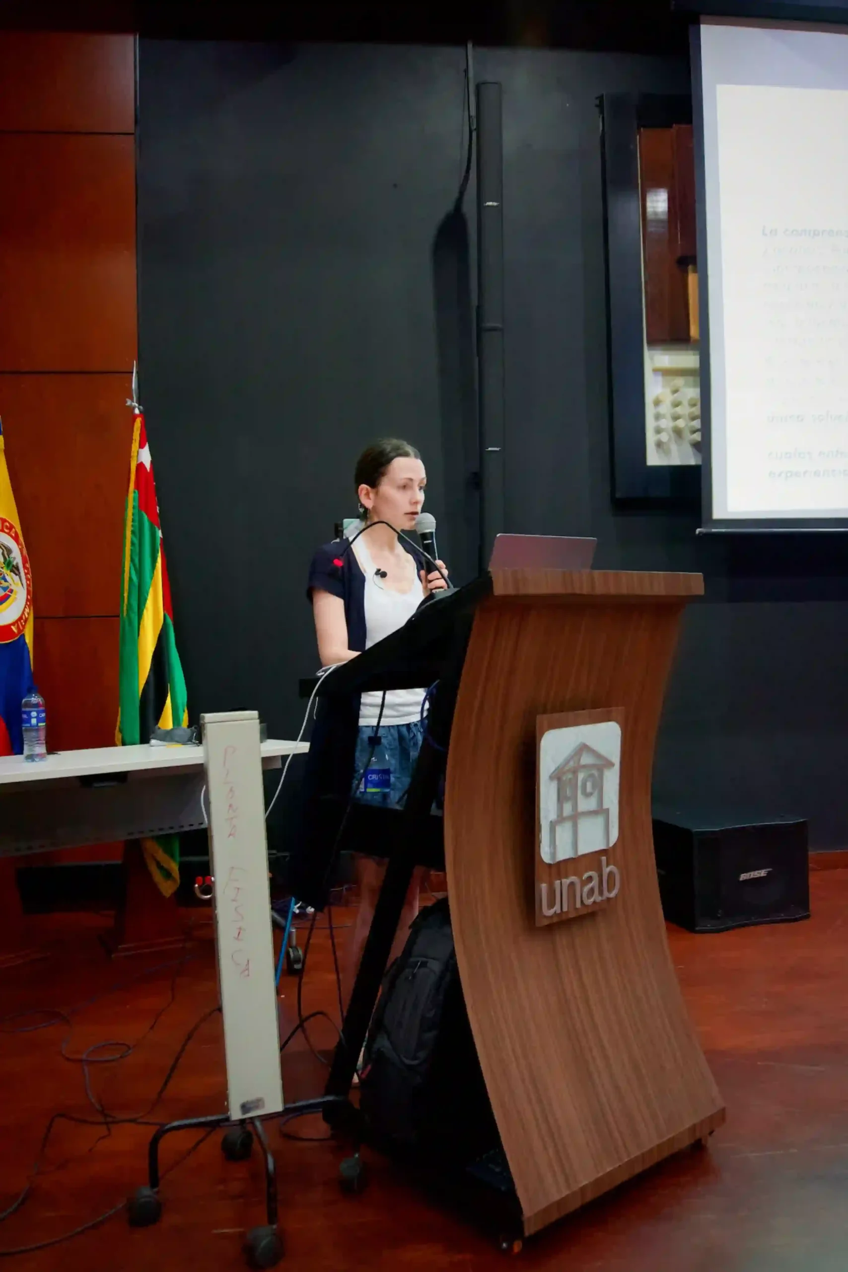 A female presenter standing by a lectern and speaking into a microphone. The walls are dark and there are flags in the corner.