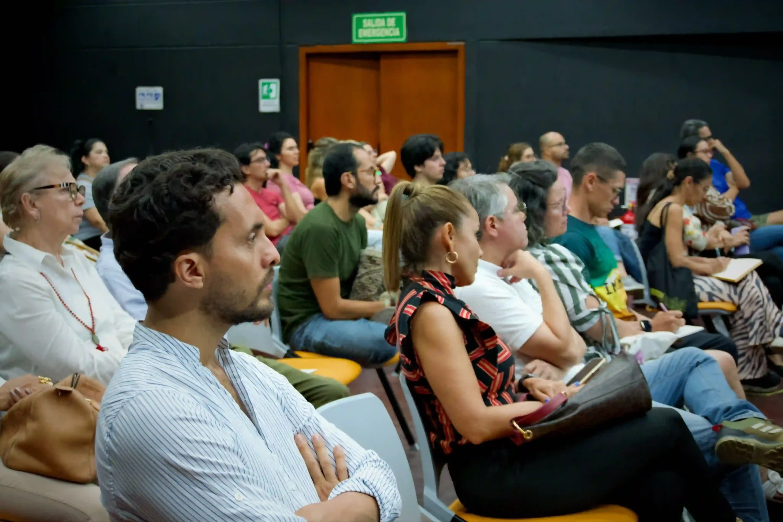 Several rows of people, 50% women and 50% men, sitting in chairs in a room with dark walls and intently listening to a presentation about body awareness and decision-making, some taking notes