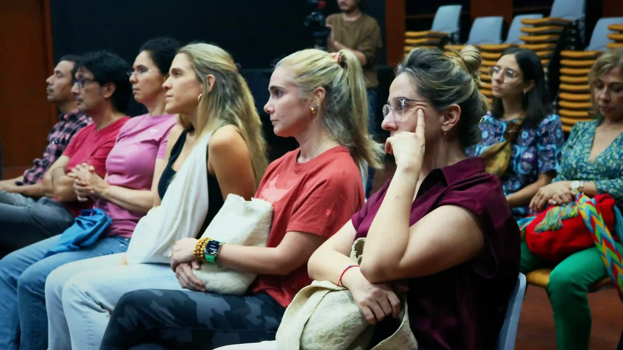 A close-up showing 2 rows of people, 60% women and 40% men, sitting in chairs and intently listening to a presentation