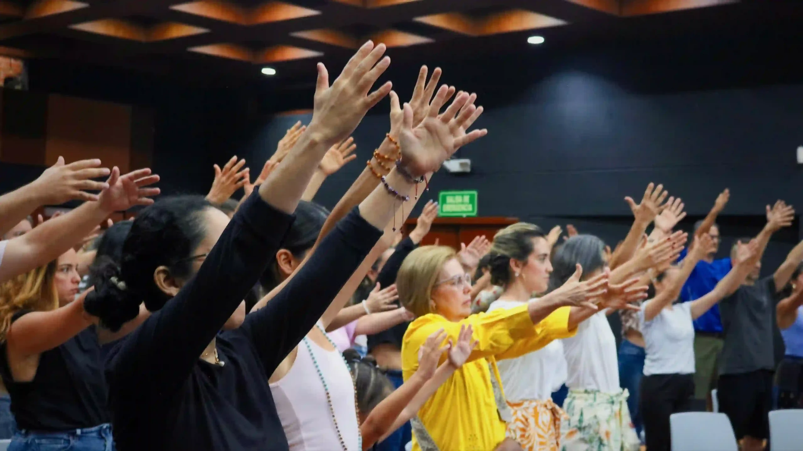 In a room with dark walls, many people a standing with their hands lifted diagonally forward, all looking in the same direction