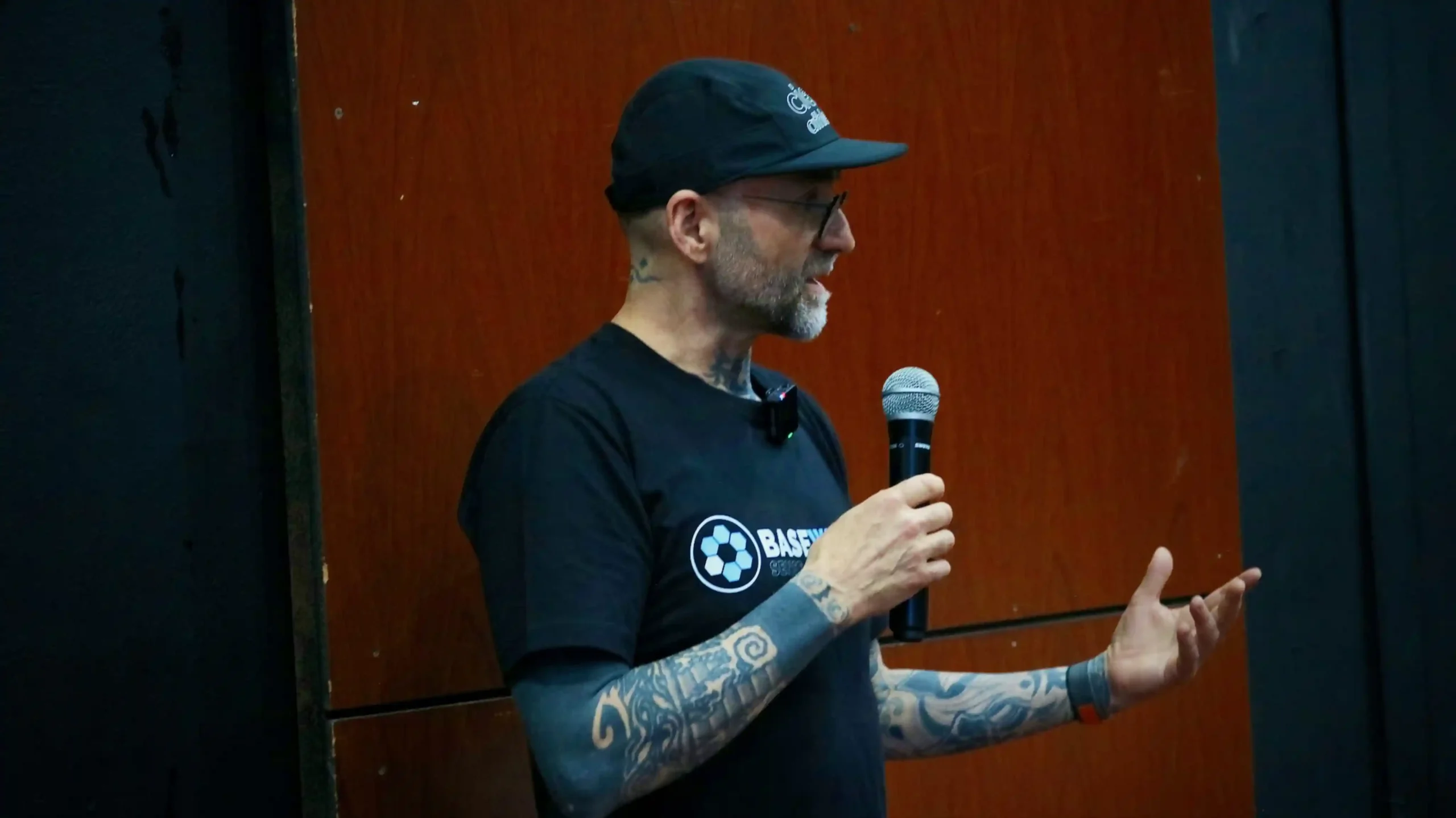 A tattooed man in glasses with a short beard wearing a black t-shirt and a black speaks into a microphone in front of a wall with wooden surface