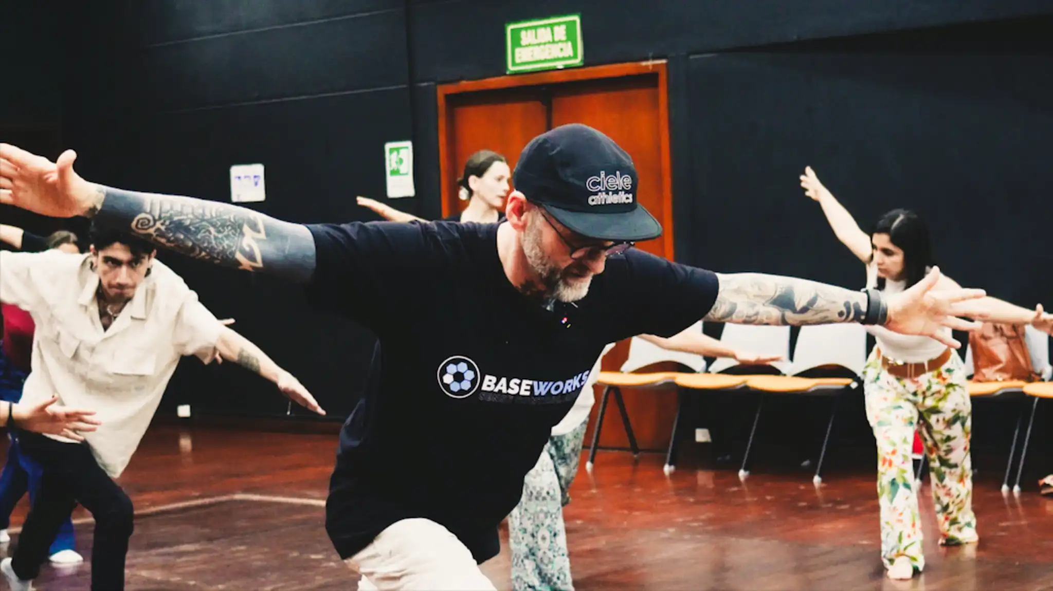 The instructor demonstrating a movement with the arms extended out to the side as he leans forward while the students mimic his movements.