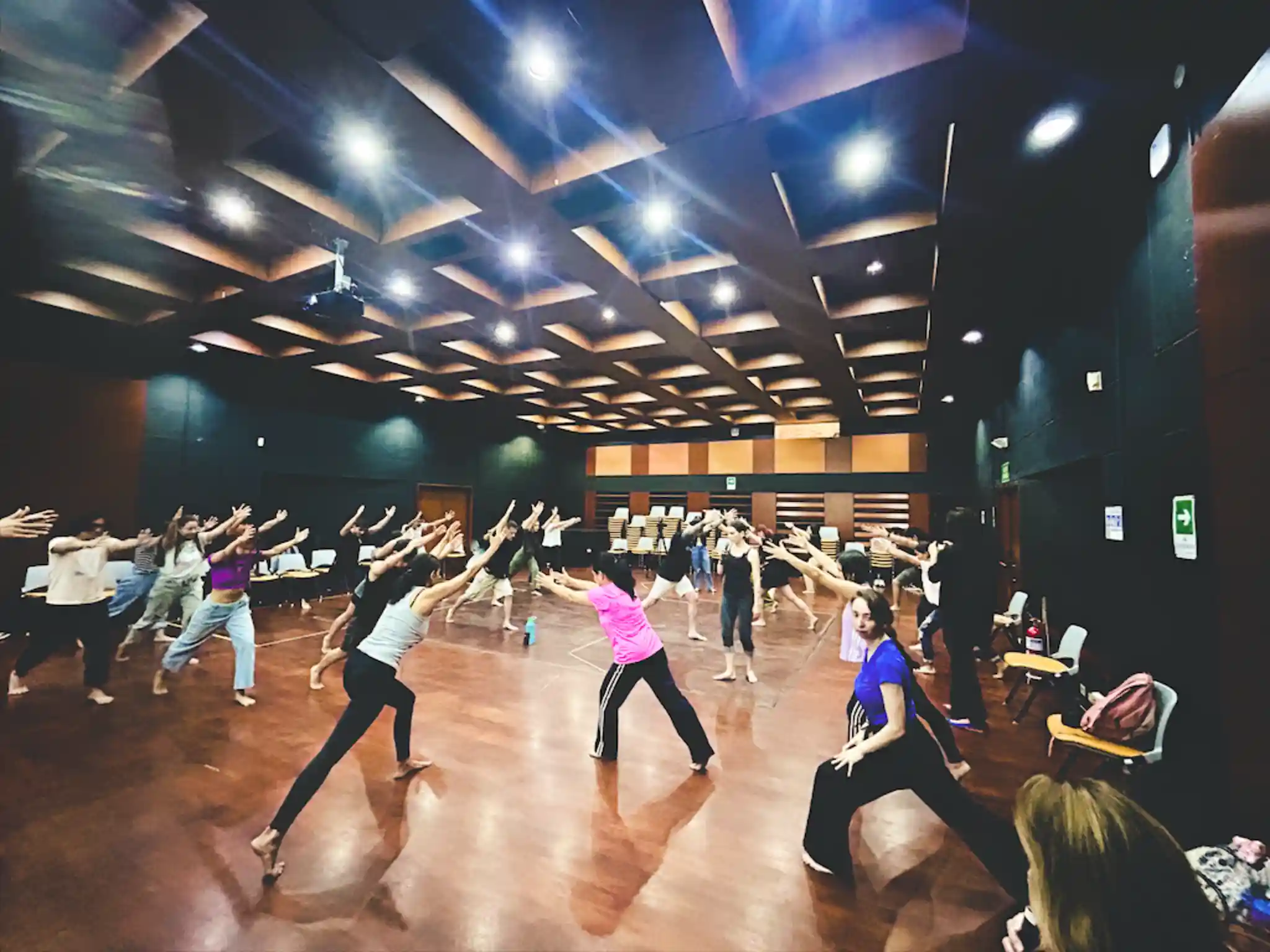 A group of students practicing a movement with the instructor demonstrating in a lunging position with their arms extended forward.