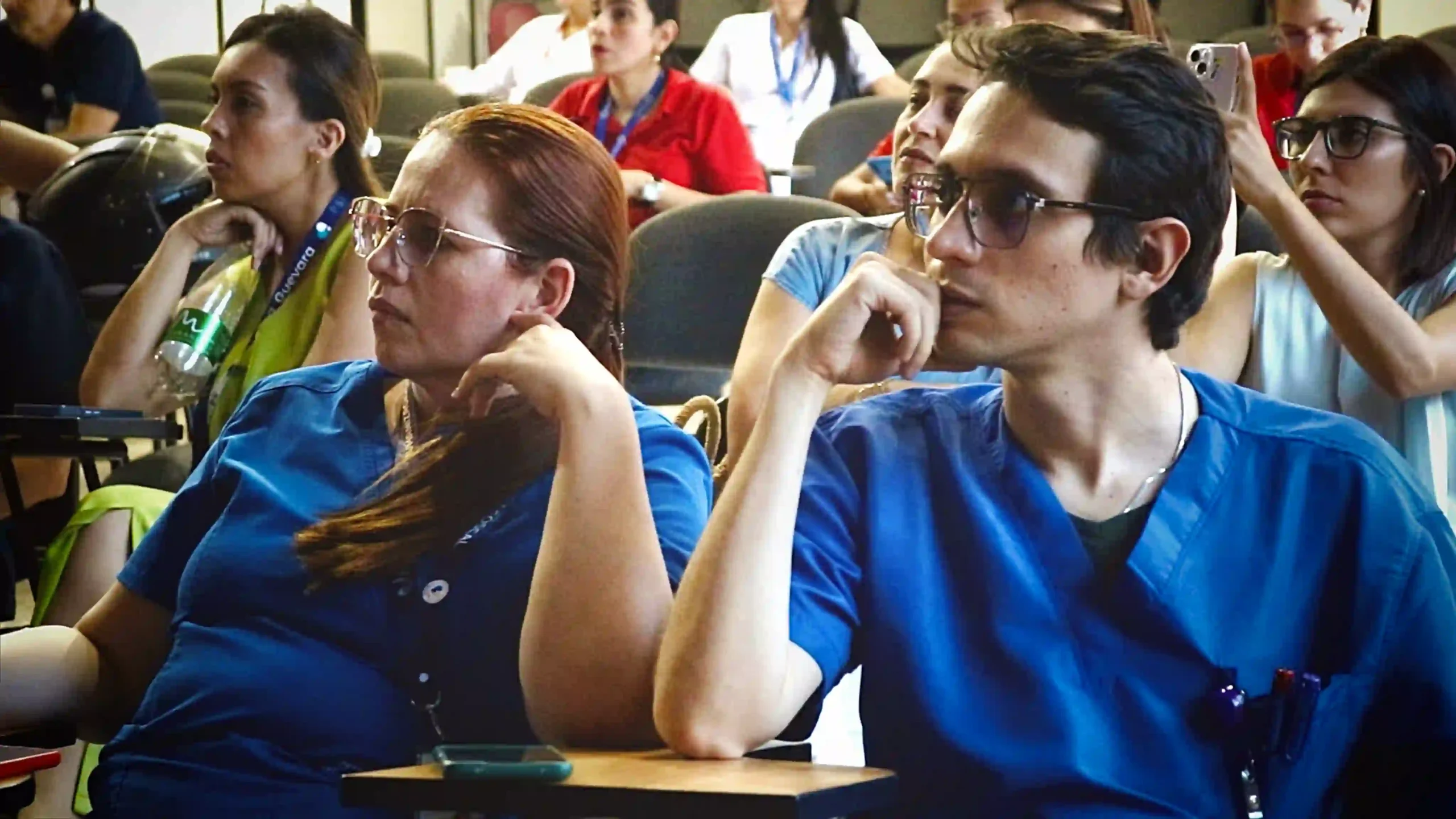 People, some of them in blue medical robes, intently listen to a presentation in a conference room with concentrated expressions, some of them are taking photos