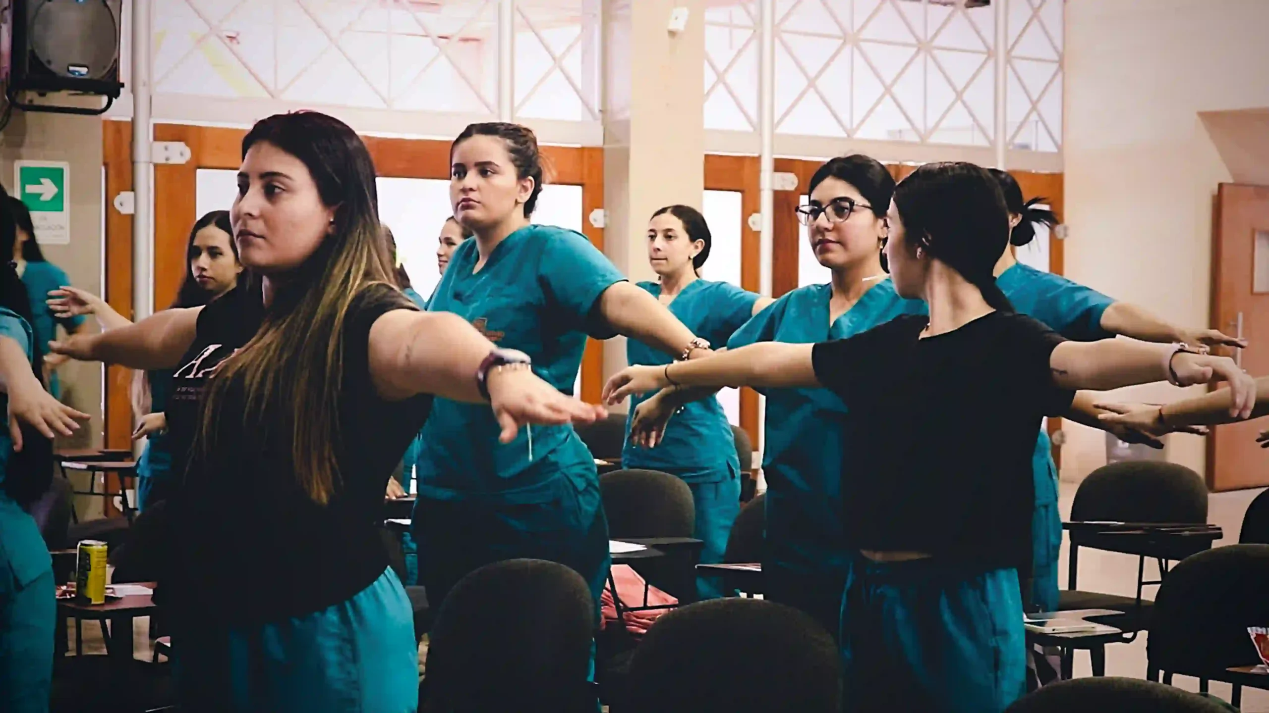 In a spacious conference room with large windows, young women wearing green medical uniforms are standing with their arms spread out, following instructions