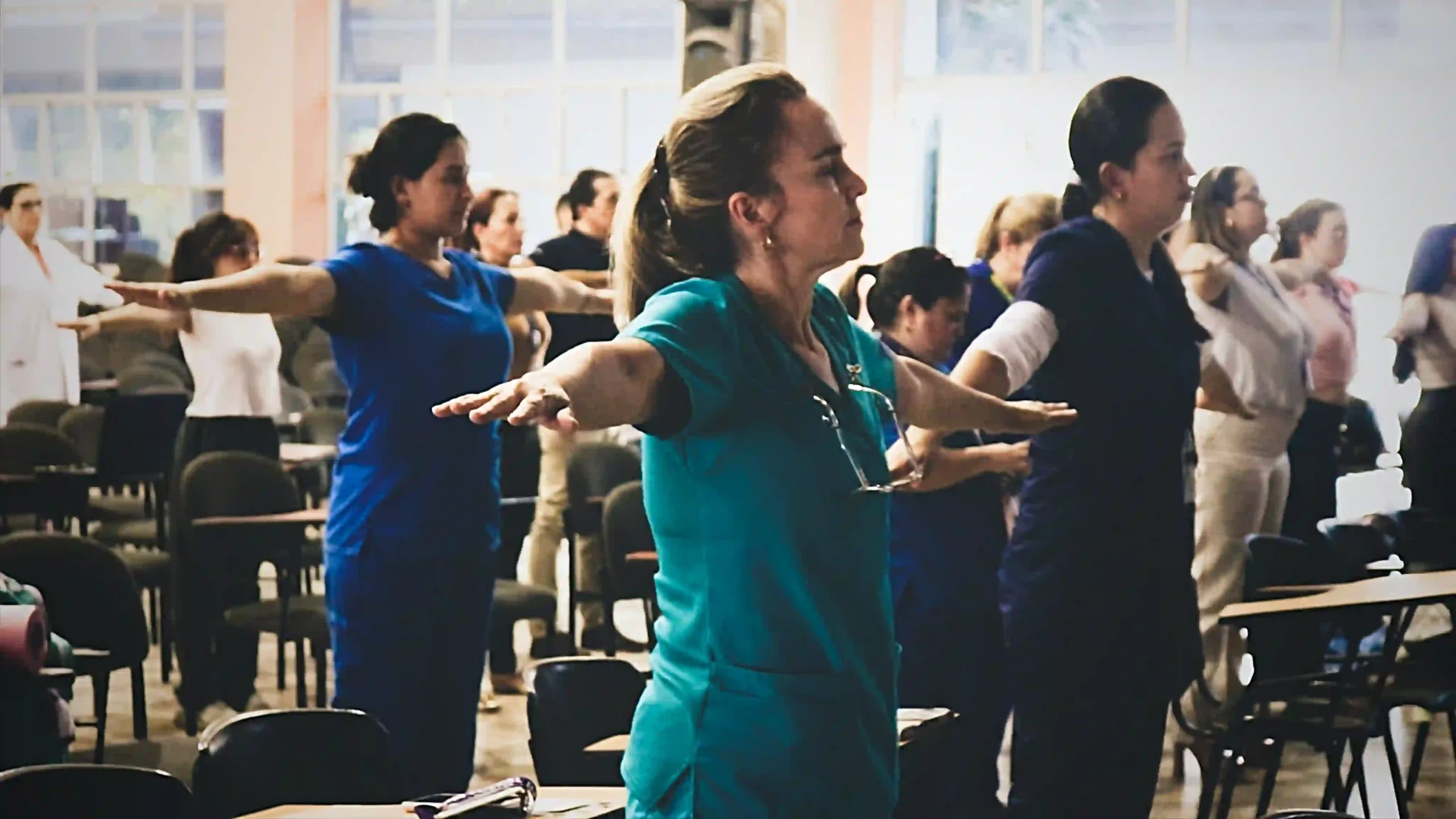 In a spacious conference room with large windows, women wearing medical uniforms are standing with their arms spread out, following instructions