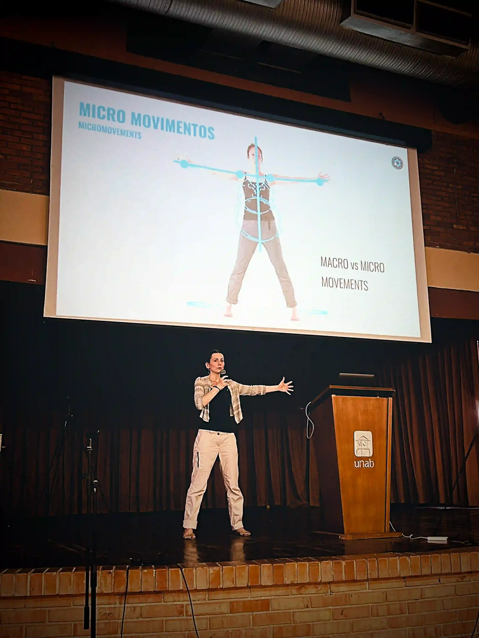 A female presenter stands on stage speaking into a microphone and spreading her other arm out. Above her, on a large screen, the slide shows a similar body position with superimposed blue symbols and the title says "MICRO MOVIMIENTIONS"