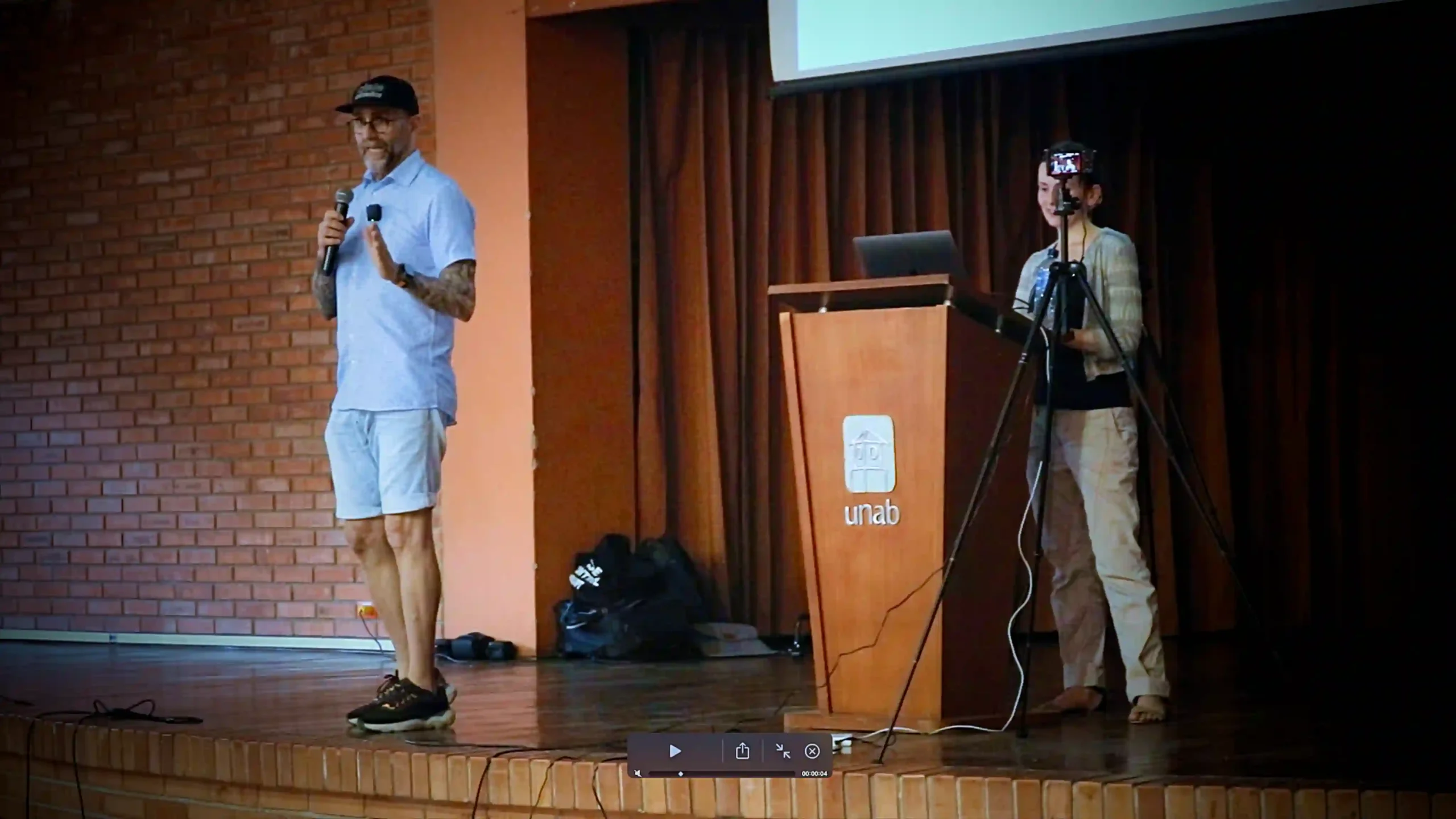 A man dressed in light T-shirt and shorts and a black cap stands on stage with a microphone explaining something and a woman stands in front of a lectern on the same stage, with a red curtain in the background