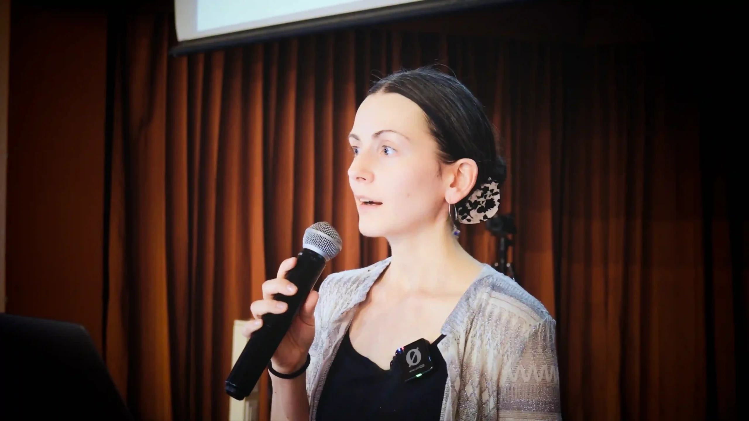 A female presenter is speaking into a microphone with a red curtain of a conference hall in the background