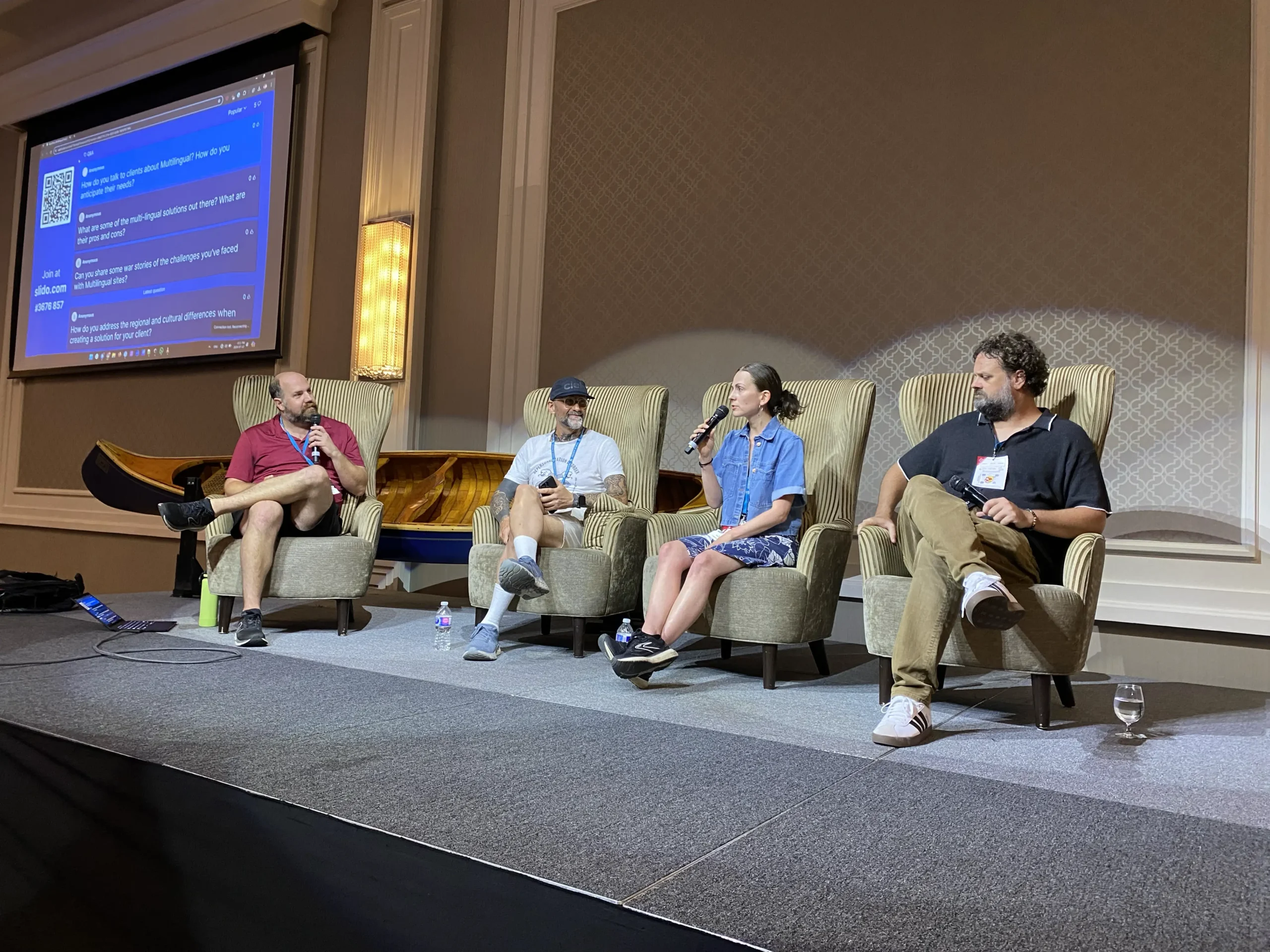 3 men and 1 woman sitting in armchairs on stage having a conversation using microphones during The WordPress Multilingual Development Panel Discussion at WordCamp Canada 2024 in Ottawa. Left to Right: Stéphane Boisvert, Patrick and Asia from Baseworks, Trevor Mills.