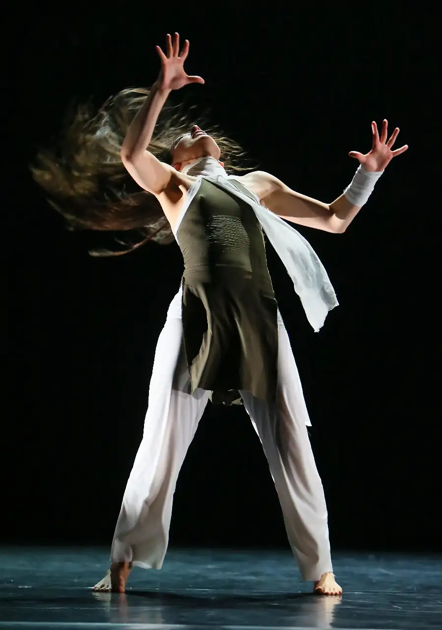 Maria Lucia is dressed in white pants and a dark green top, captured mid-dance, expressing fluidity in her motion as a result of her education through dance