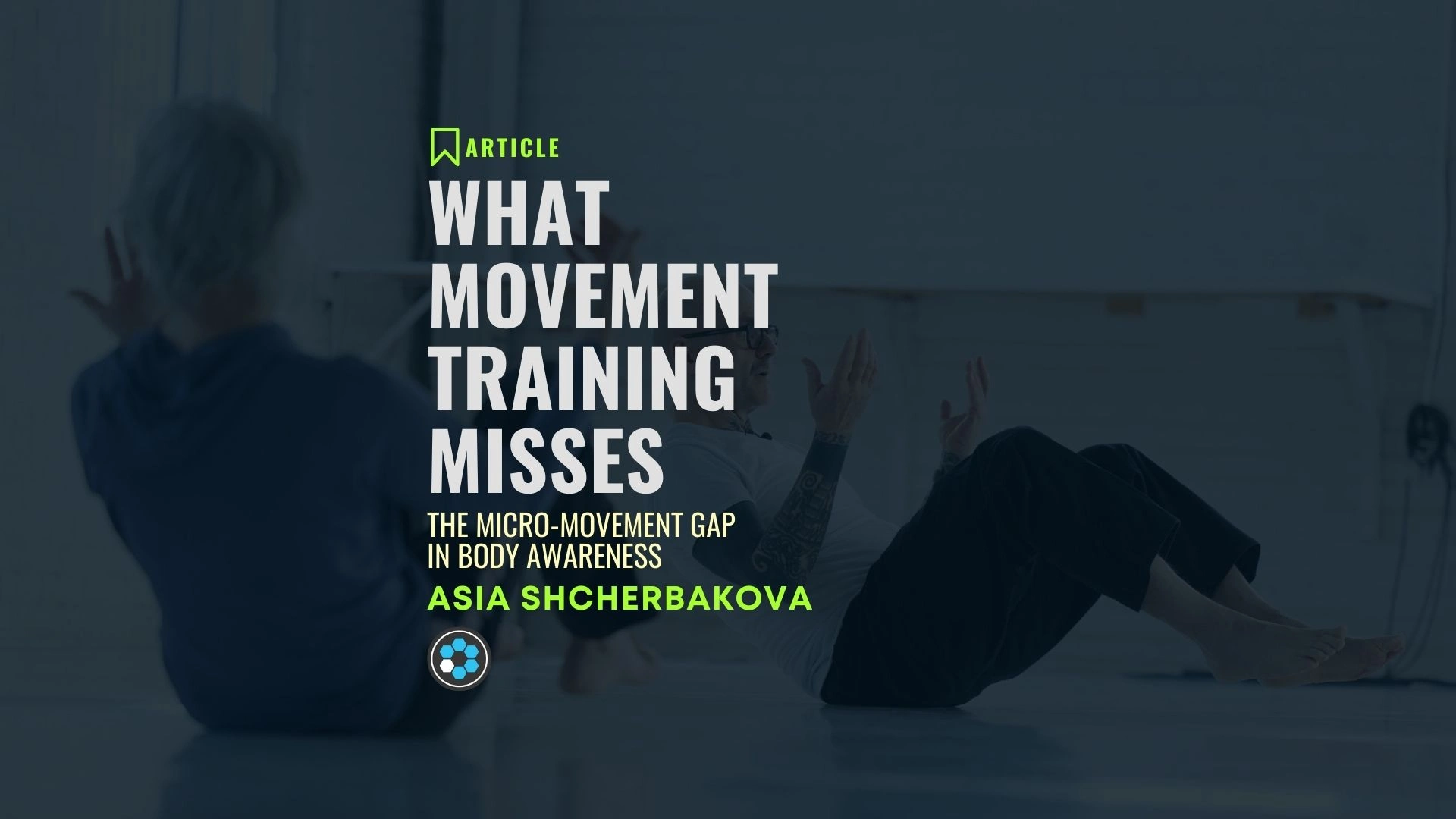 Two individuals seated on the floor doing slow, controlled movement exercises in a spacious studio.