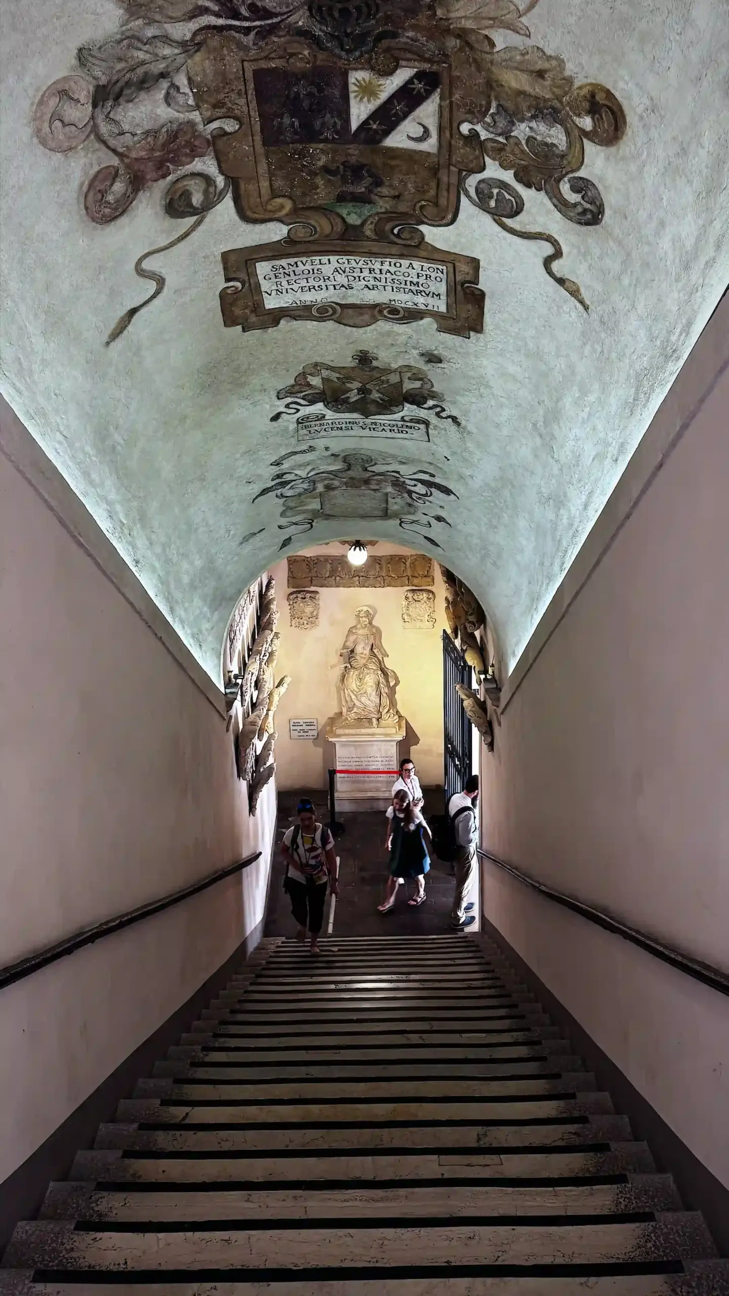 Looking up a long barrel-vaulted staircase at Palazzo del Bo. Fresco paintings and Latin inscriptions cover the vaulted ceiling. Conference participants descend the stairs, with a classical statue visible at the lower landing.