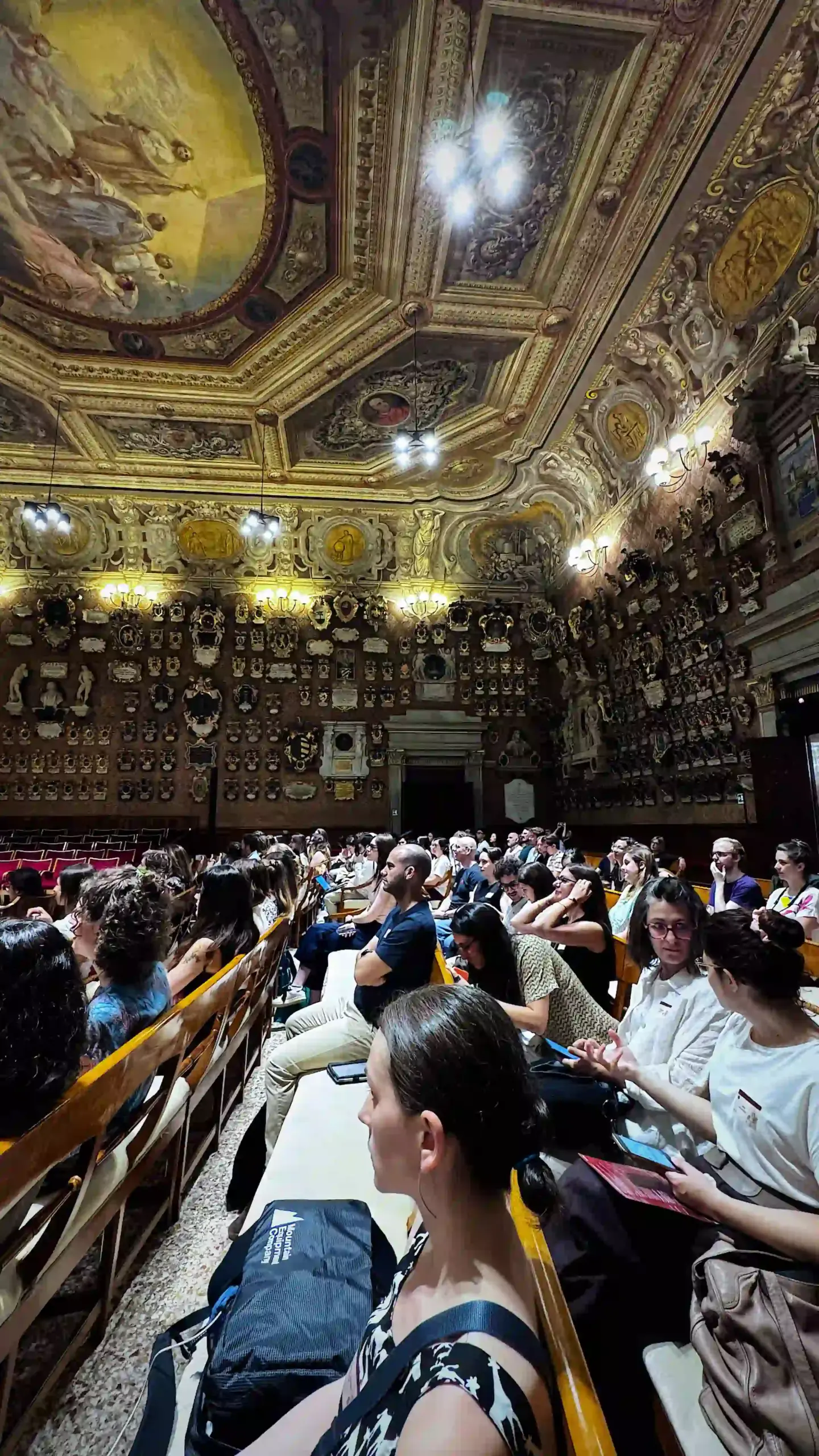 A packed audience seated in wooden pews in the Aula Magna Galileo Galilei of Palazzo del Bo, viewed from the upper rows. The elaborate baroque ceiling features gold-painted frescoes and painted medallions. Carved heraldic shields line the walls from floor to cornice.