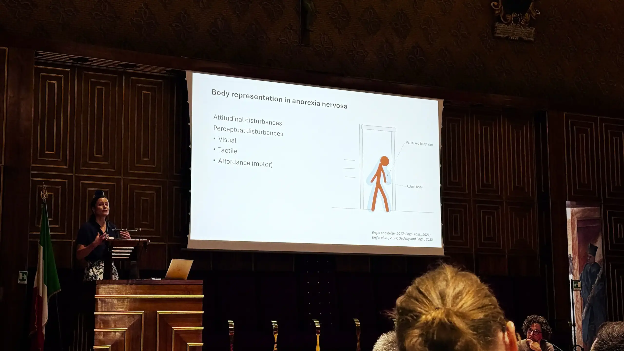 A researcher presents at the podium in the Aula Magna Galileo Galilei. The projected slide is titled "Body representation in anorexia nervosa" and lists attitudinal and perceptual disturbances alongside a diagram comparing perceived versus actual body size. An Italian flag stands to the left of the podium.