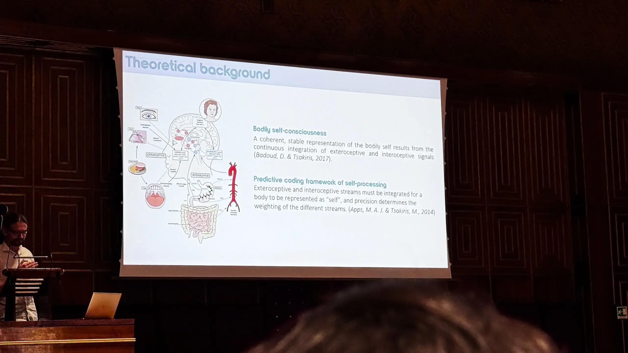 A researcher presents at the podium in the Aula Magna Galileo Galilei. The slide titled "Theoretical background" displays a diagram of exteroceptive and interoceptive sensory integration, alongside text defining bodily self-consciousness and the predictive coding framework of self-processing, citing Badoud and Tsakiris (2017) and Apps and Tsakiris (2014).