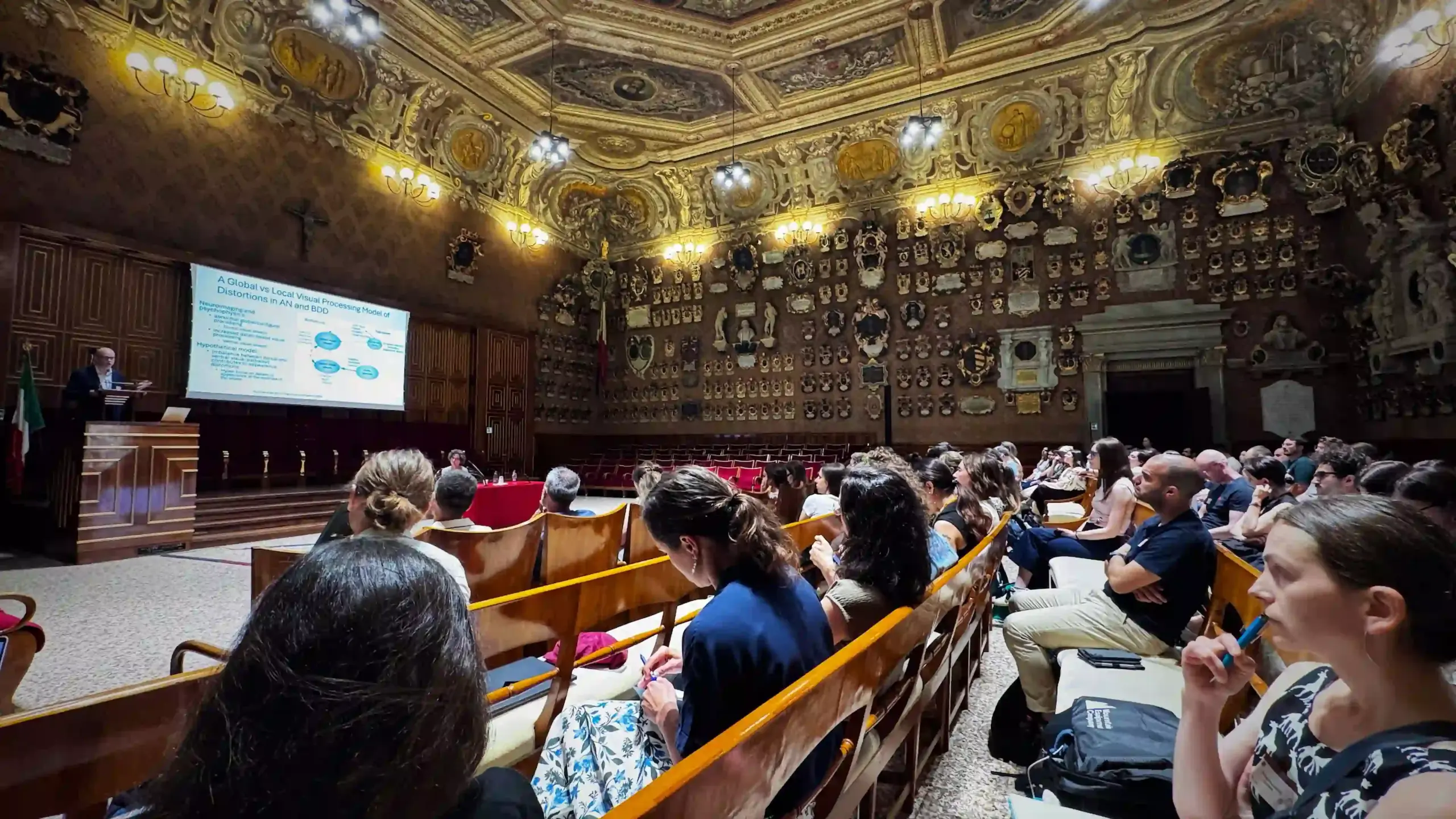 Wide-angle view of the Aula Magna Galileo Galilei of Palazzo del Bo during a conference talk. Attendees fill the wooden pews. A projected slide at the far end reads "A Global vs Local Visual Processing Model of Distortions in AN and BDD." The ornate baroque ceiling and heraldic-lined walls are fully visible from this angle.