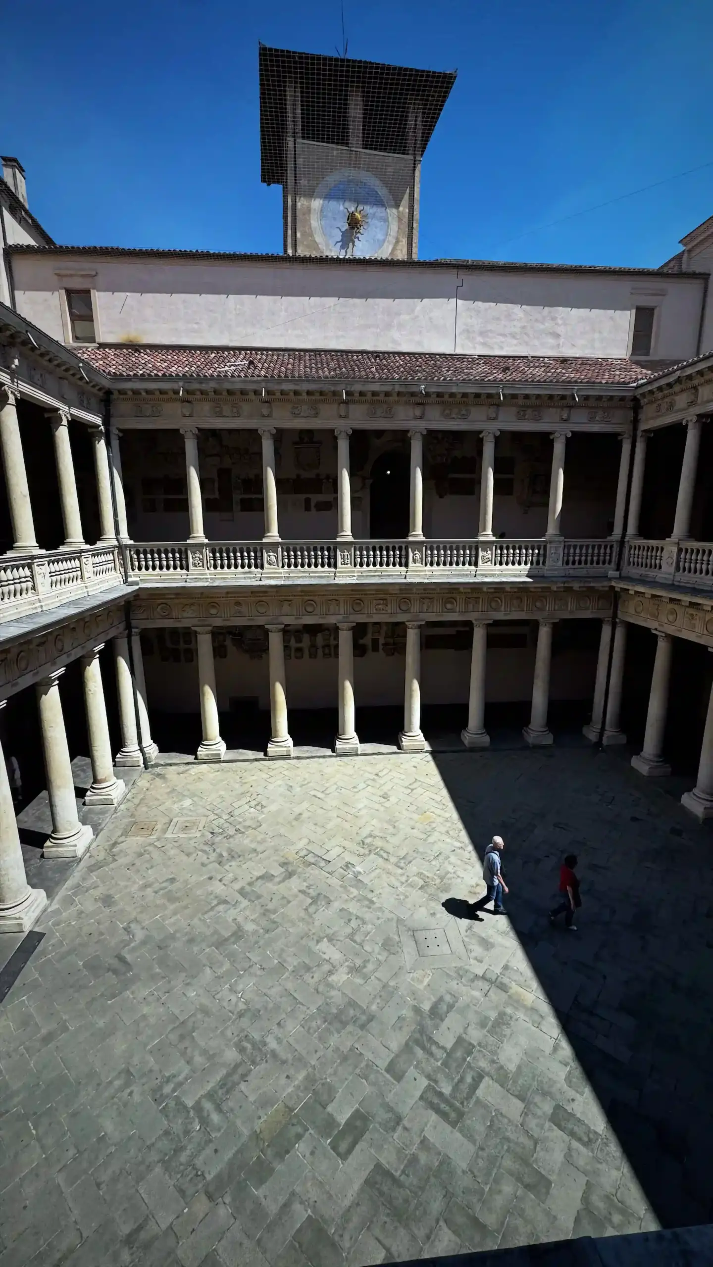 Looking down into the Renaissance inner courtyard of Palazzo del Bo from an upper floor. Two tiers of colonnaded arcades with stone balustrades line all four sides of the open rectangular courtyard. A clock tower bearing the university's bull emblem rises above the roofline against a clear blue sky. Two figures cross the stone-paved courtyard below.
