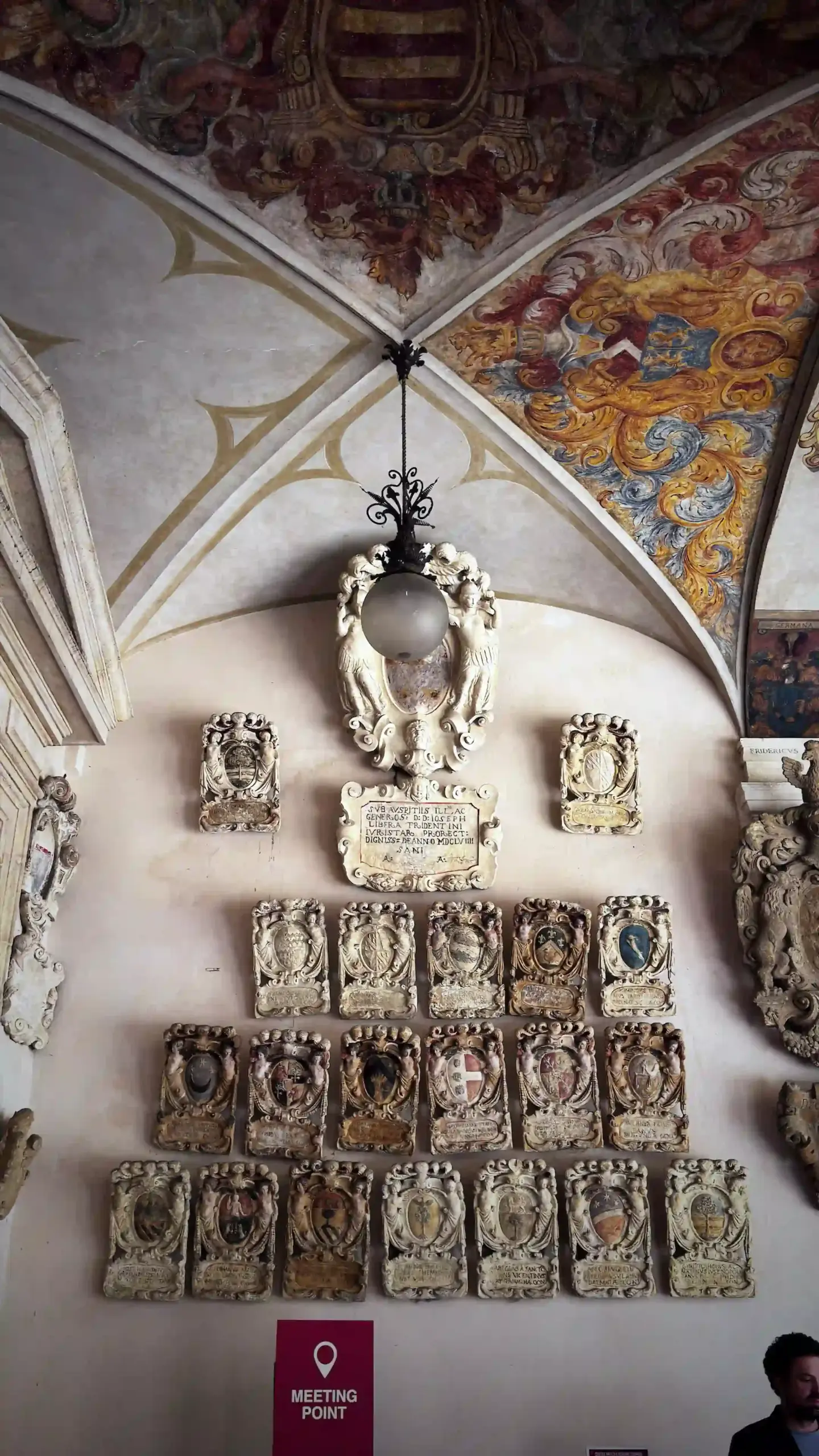 A vaulted corridor at Palazzo del Bo viewed looking up and along the wall. Rows of ornate stone escutcheons, heraldic plaques bearing the carved coats of arms of past students and rectors, cover the walls from floor to vaulted ceiling. A red "Meeting Point" sign is visible at the bottom of the frame.