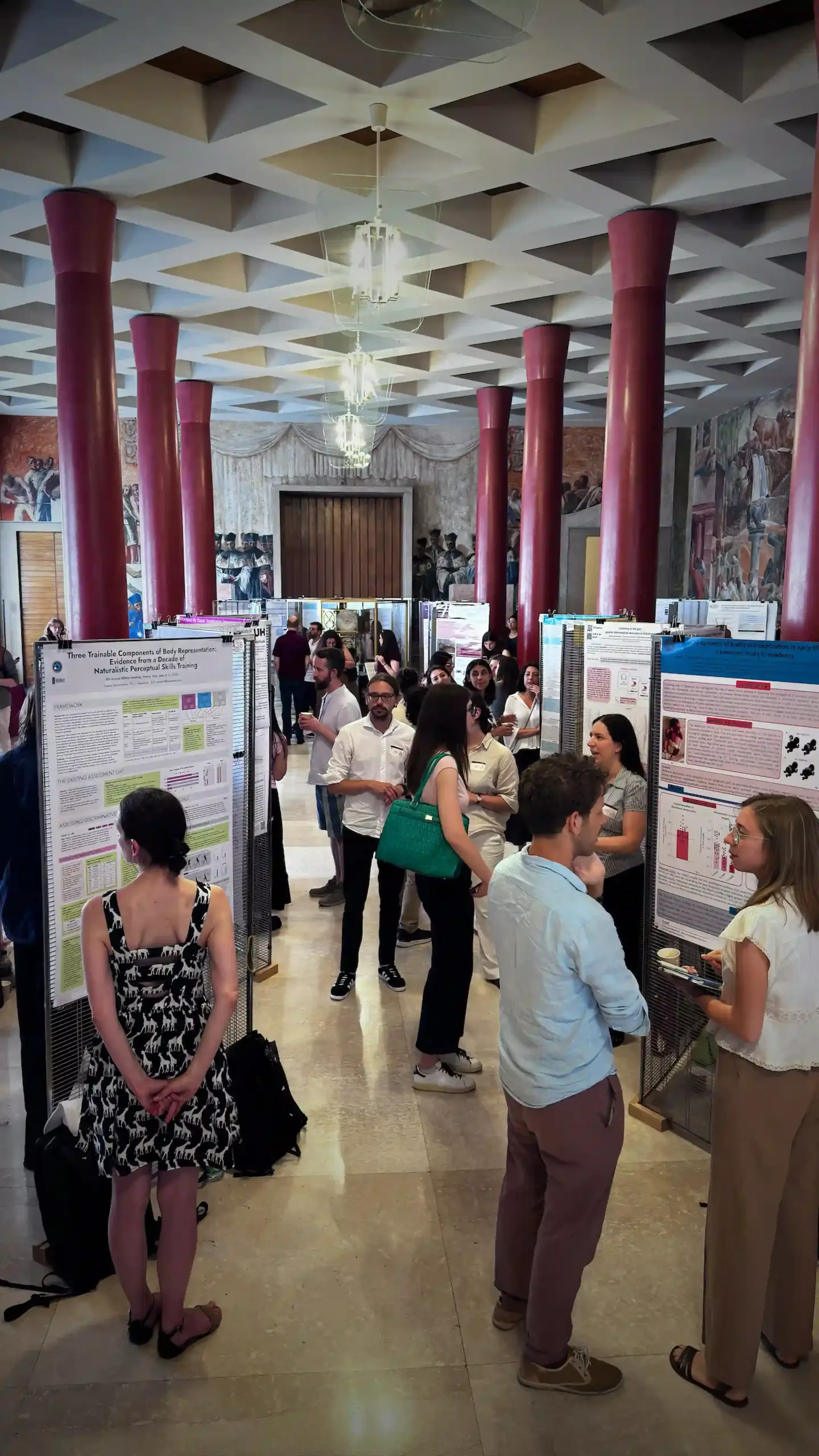 The BRNet 2026 poster session in a mid-century hall with tall magenta-red columns and decorative wall frescoes. Rows of research posters on mesh stands fill the room. The Baseworks poster "Three Trainable Components of Body Representation" is visible at the far left. Researchers and attendees circulate between the posters.