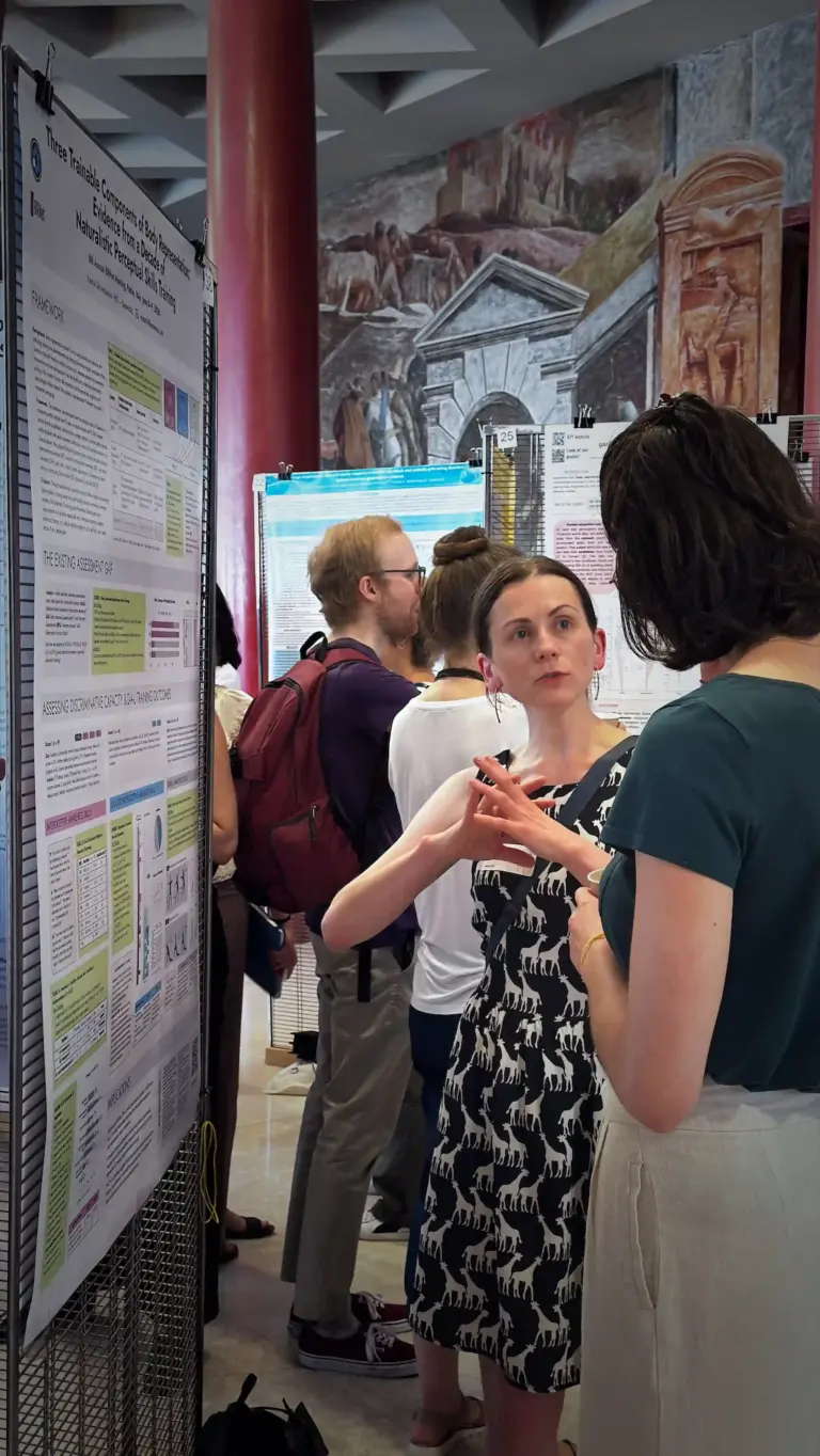 Ksenia Shcherbakova stands beside the Baseworks research poster on a mesh display stand, in conversation with another conference participant. Additional posters and attendees are visible in the background of the poster session hall.