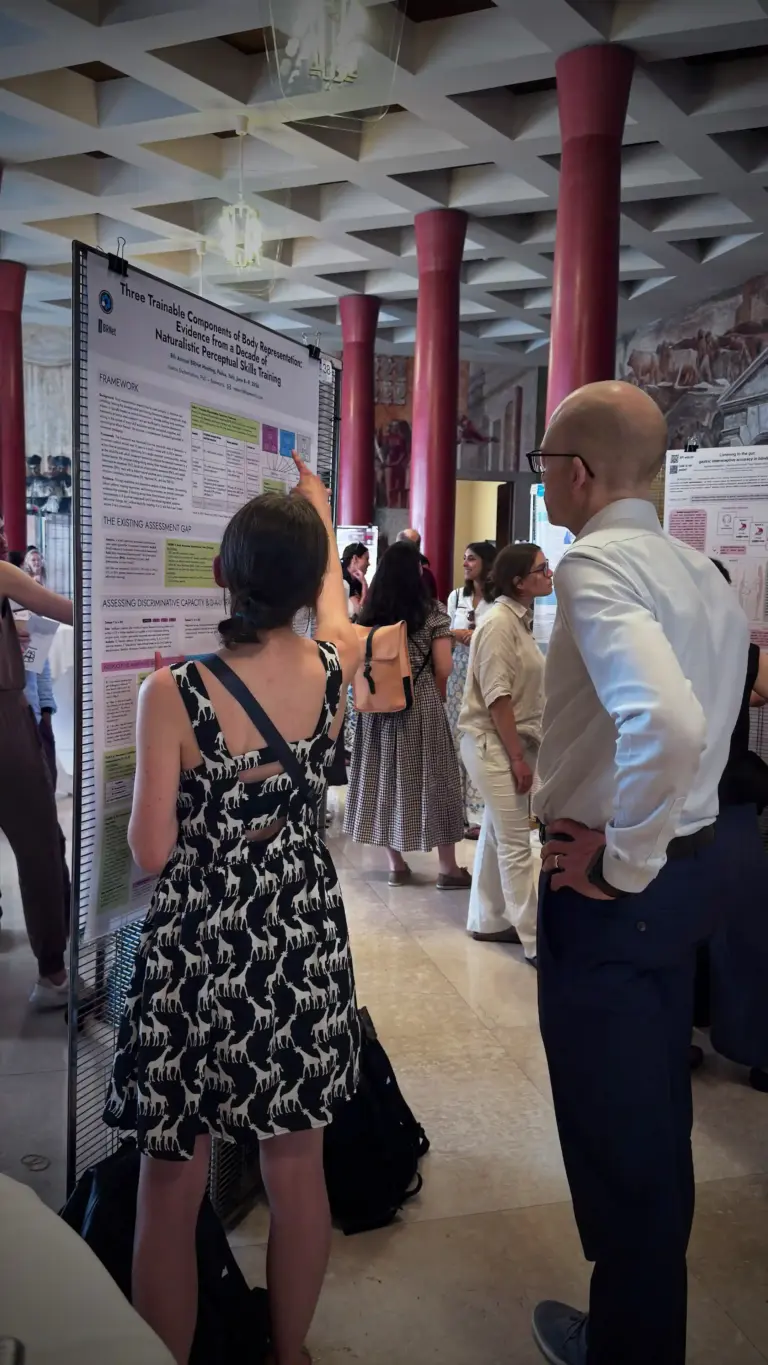 Ksenia Shcherbakova stands beside the Baseworks research poster and points to a specific section, explaining it to a visitor. The poster title "Three Trainable Components of Body Representation: Evidence from a Decade of Naturalistic Perceptual Skills Training" is legible at the top.