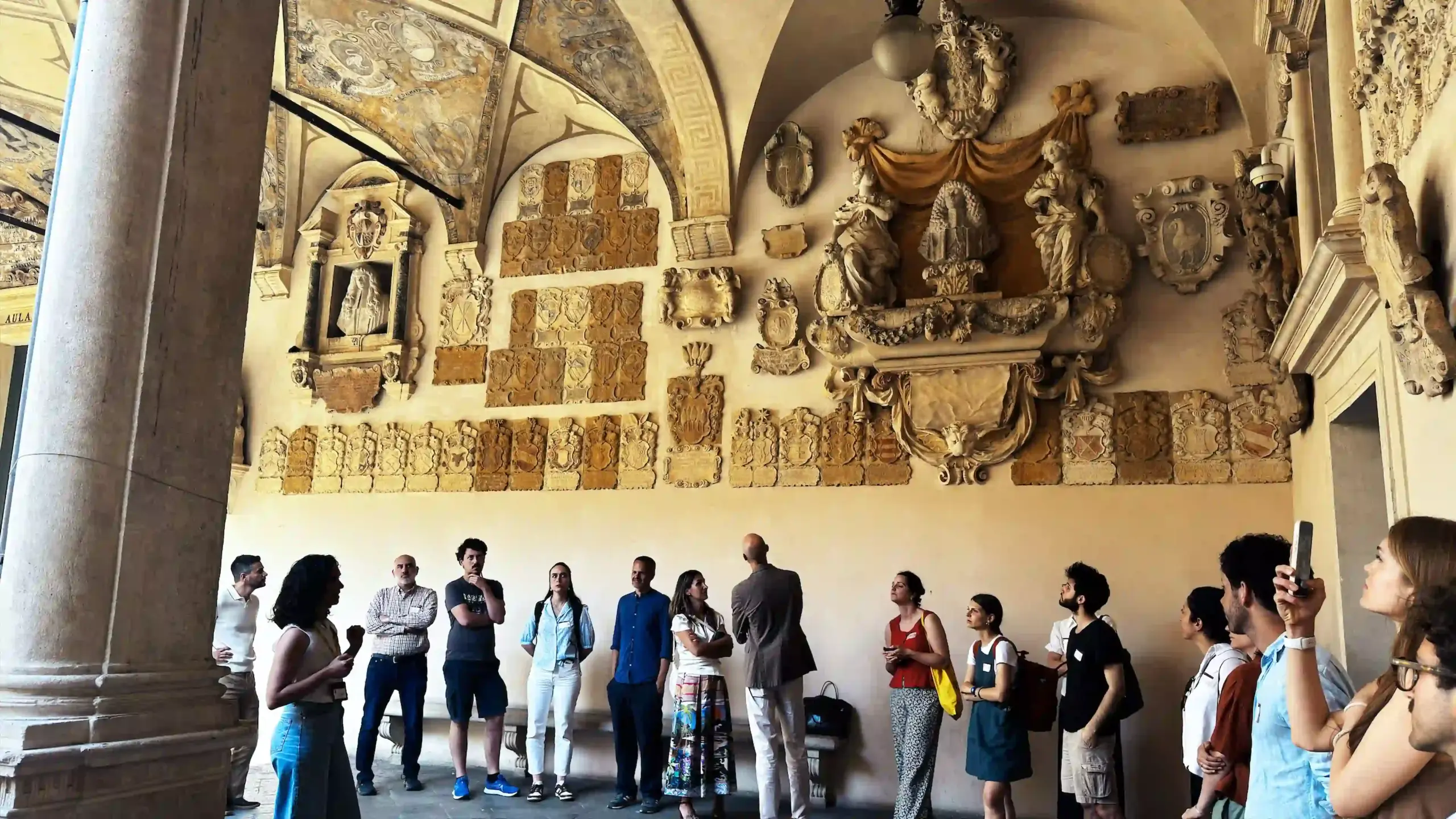 A group of conference participants stands in the ground-floor loggia of Palazzo del Bo, looking upward at the rows of ornate stone heraldic escutcheons that cover the arched wall. Several participants photograph the plaques. Classic stone columns frame the foreground.