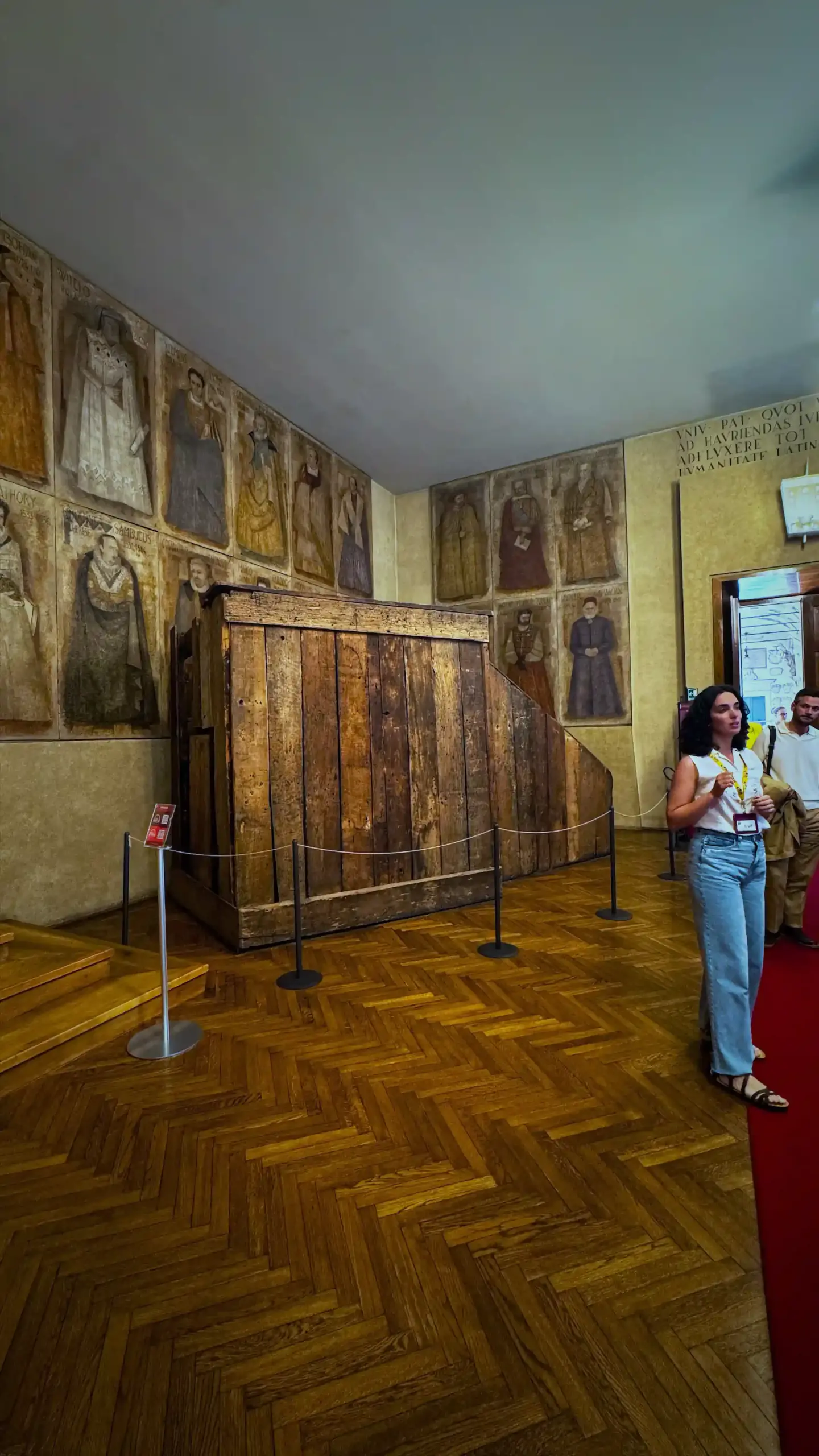 A historic ceremonial room at Palazzo del Bo. A large original wooden raised lecturer's cathedra, the traditional professor's elevated pulpit, stands roped off in the centre of the room. The walls are lined with painted portraits of past rectors and professors in period dress.