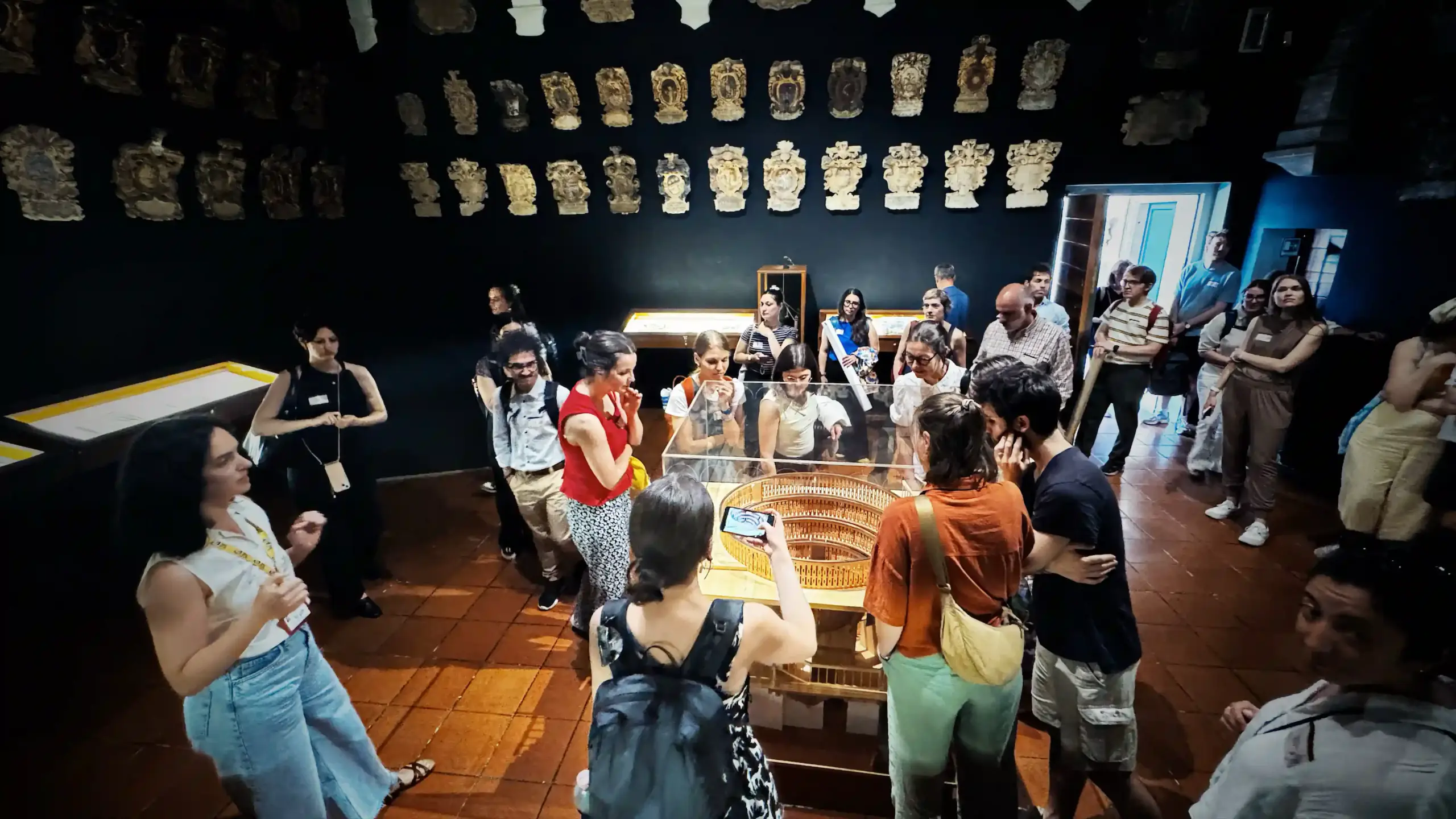 BRNet 2026 participants gathered in a circle around a glass display case containing a detailed wooden scale model of the Teatro Anatomico of Padova. The room has dark-painted walls displaying rows of white plaster heraldic reliefs above. Participants lean in to look at the model.