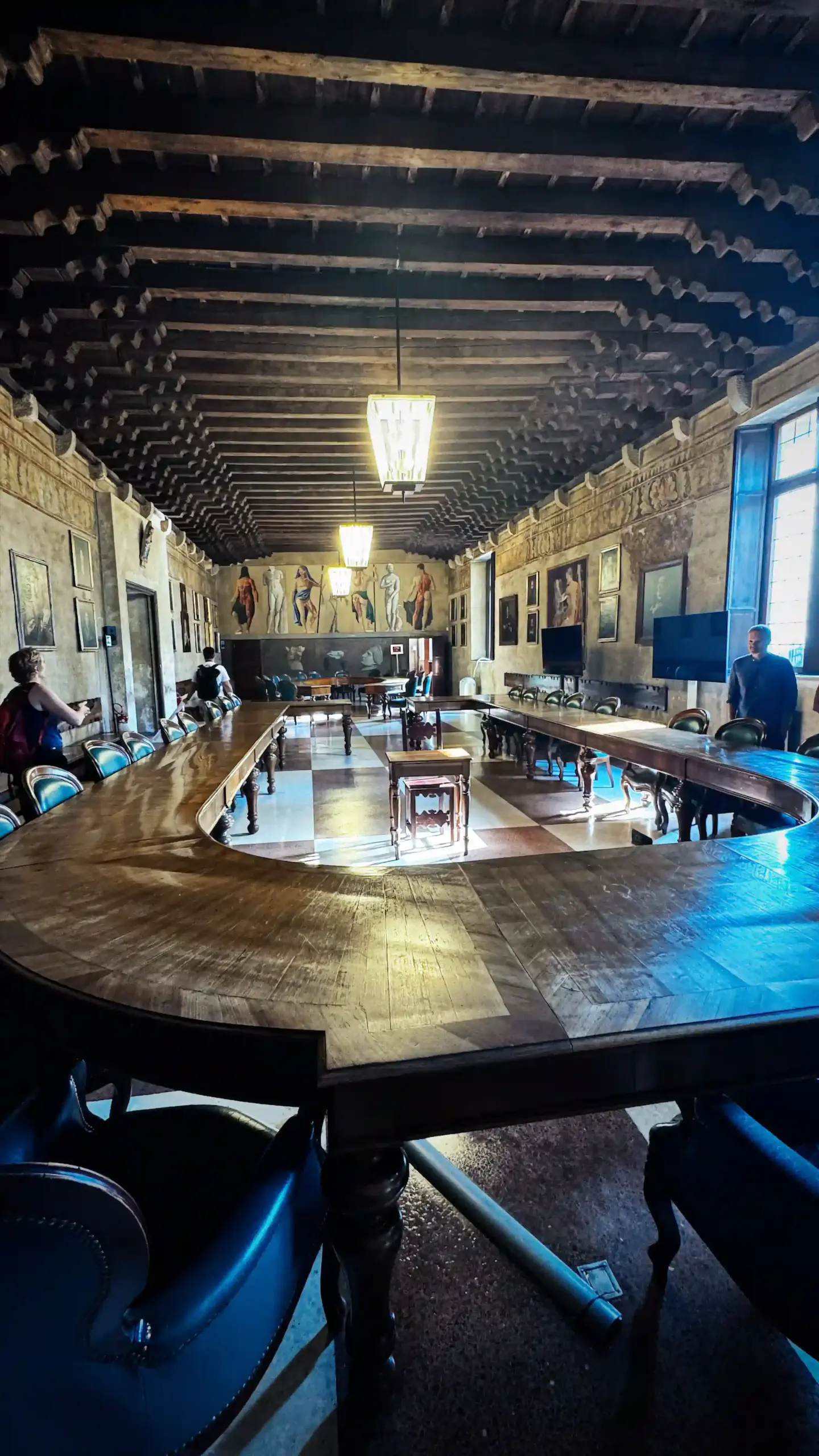 A large ceremonial council room at Palazzo del Bo. A horseshoe-shaped wooden table with matching leather chairs fills the centre of the room. The ceiling has exposed dark wooden beams, and frescoed walls depict classical human figures. Two windows let in natural light from the right side.