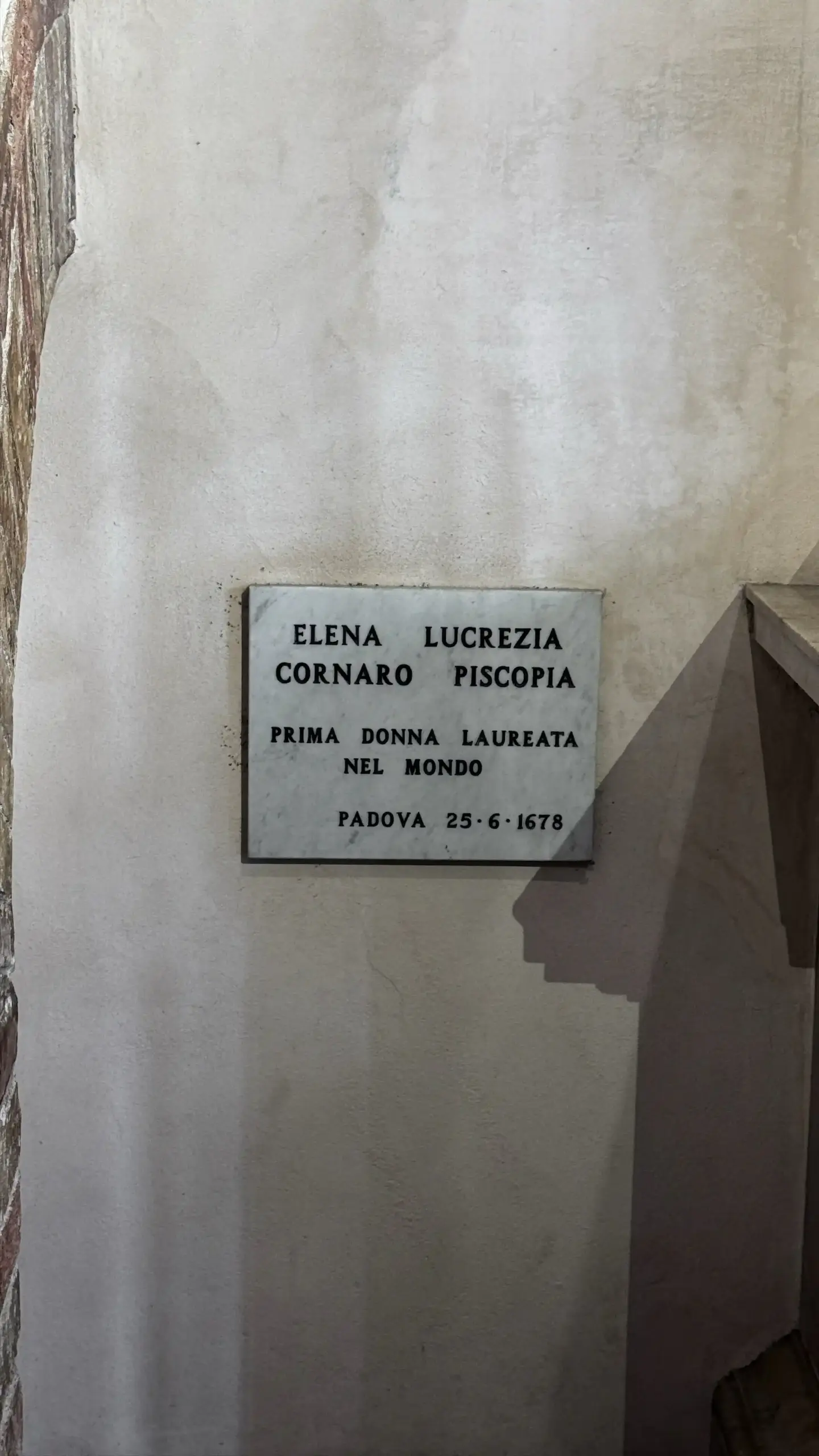 A marble commemorative plaque mounted on a rough stone wall. The inscription reads: Elena Lucrezia Cornaro Piscopia, Prima Donna Laureata nel Mondo, Padova 25-6-1678.