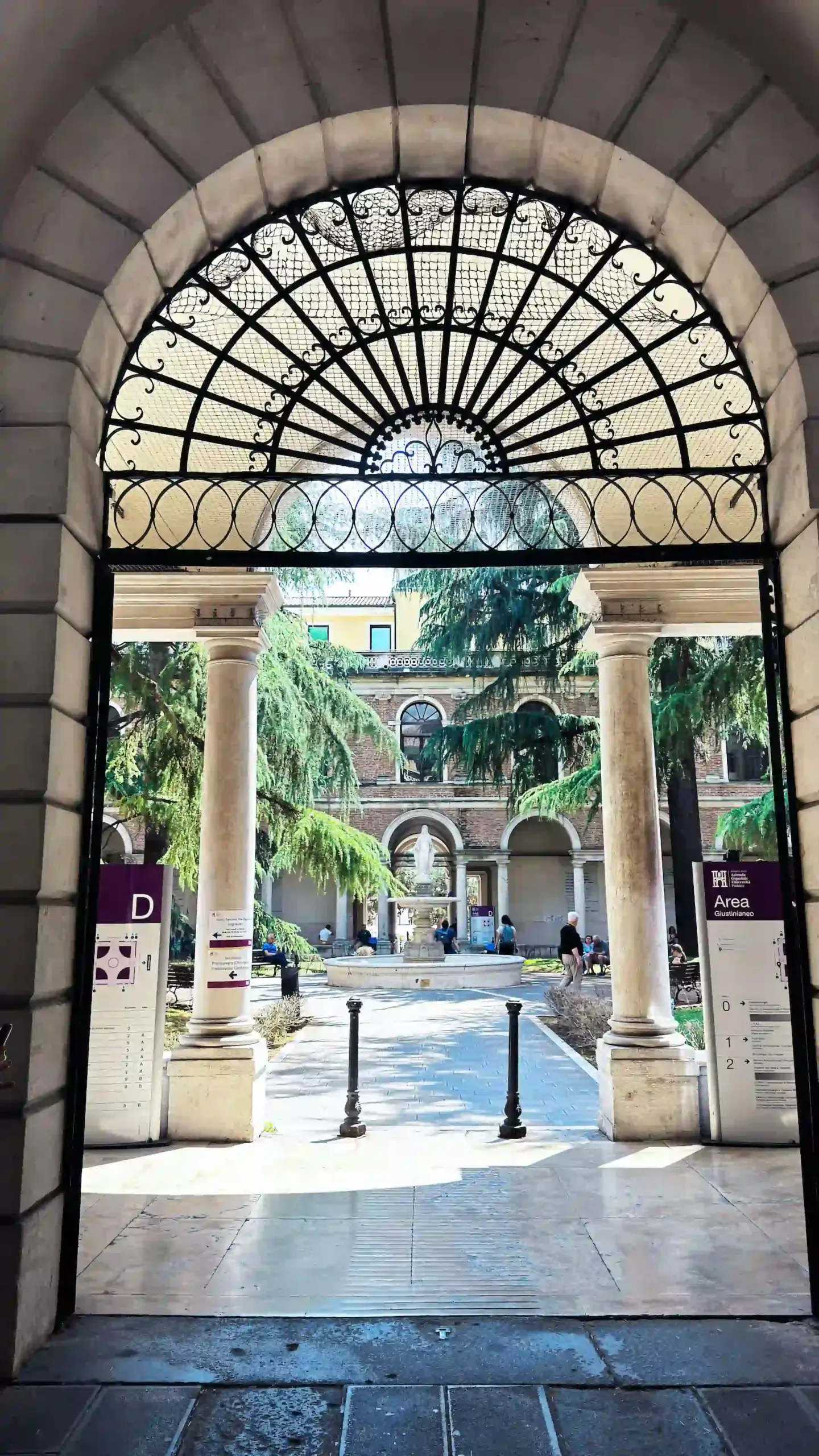 View through an ornate semicircular iron-lattice archway into the courtyard of the Area Giustinianea, University of Padova. Beyond the open gate, a classical arcade with stone columns borders the courtyard, planted with tall trees and leading to a Renaissance building in the background. University of Padova directional signs are mounted to each side of the entrance. The Area Giustinianea is the University of Padova's medical faculty and hospital complex; it hosted Day 2 of BRNet 2026, with the conference auditorium situated within the complex.