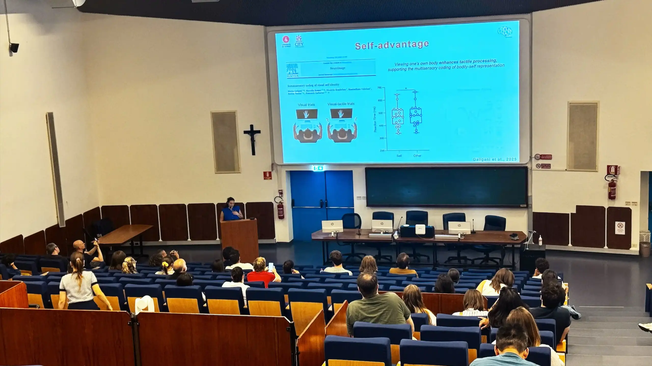 A presenter at the podium in the lecture auditorium at BRNet 2026, Day 2. The projected blue slide reads Self-advantage and includes experimental data diagrams. Audience members are seated in the tiered blue seating rows. Area Giustinianea, University of Padova.