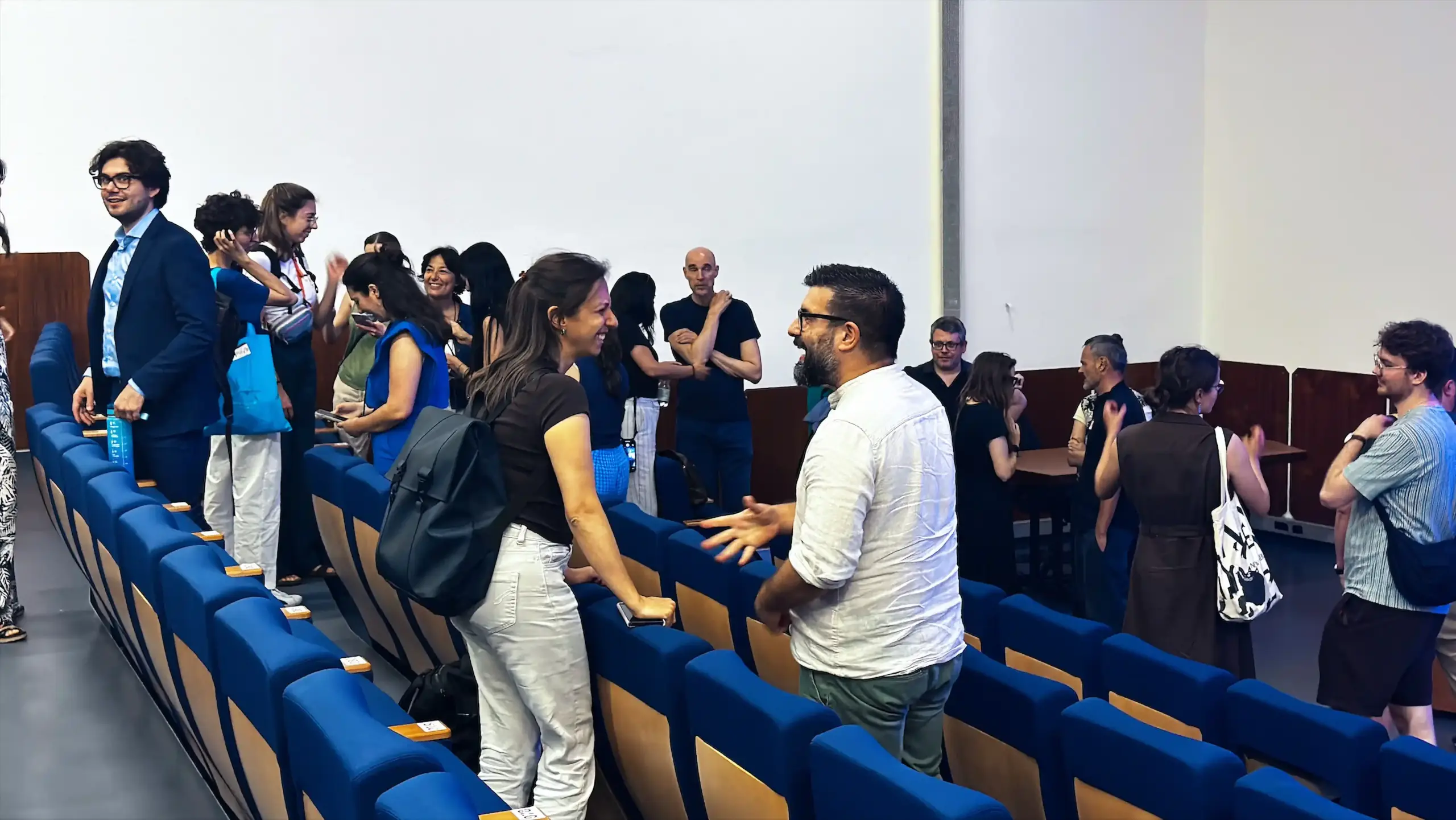 Conference attendees stand and converse in small groups in the lecture auditorium aisles during a break between sessions at BRNet 2026. Blue upholstered seating visible in the foreground. Area Giustinianea, Day 2, June 9, 2026.