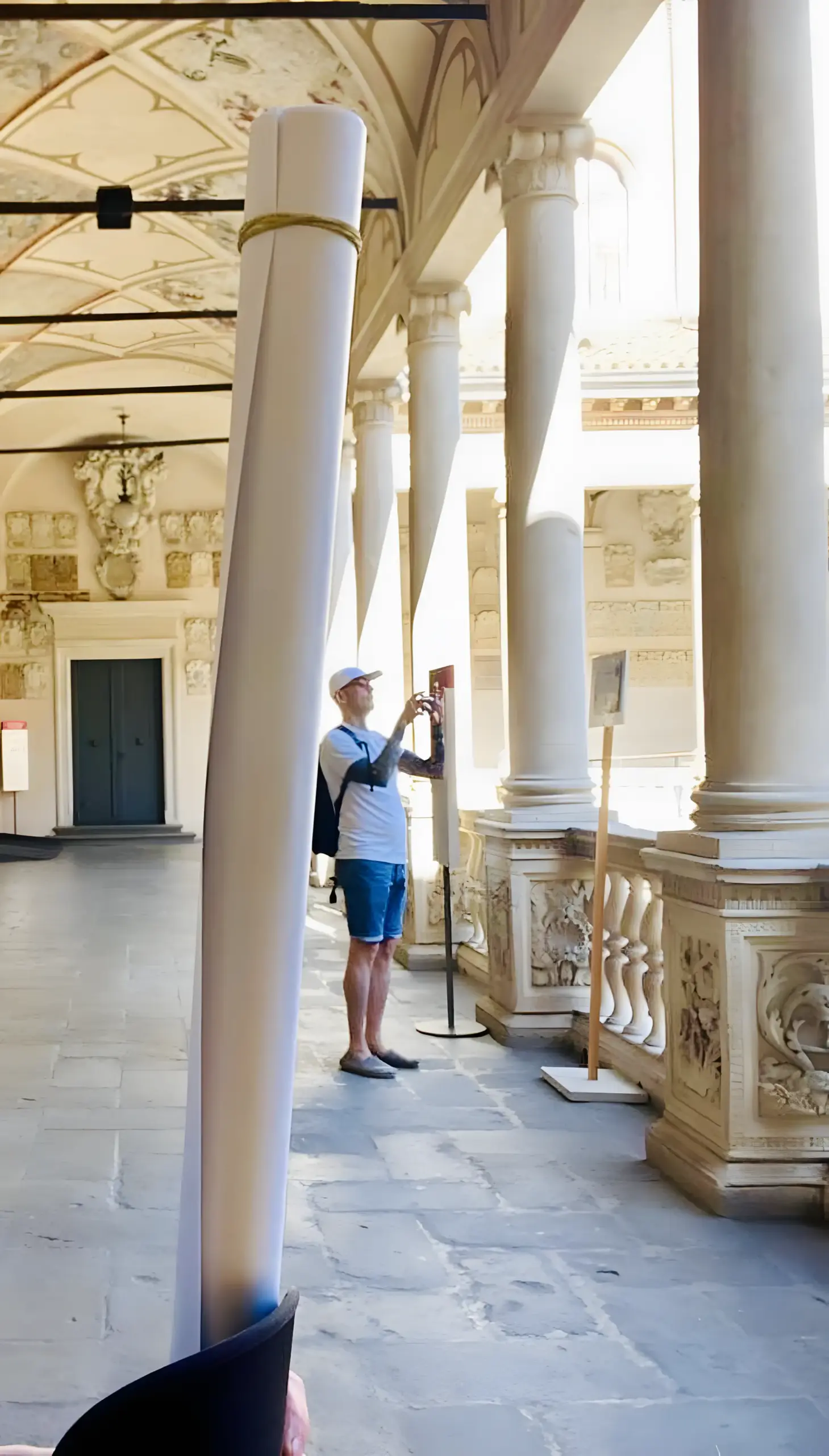 The colonnaded arcade of the University of Padova's historic courtyard. Patrick photographs the ornate columns and vaulted ceiling while Asia holds a rolled conference poster in the foreground.