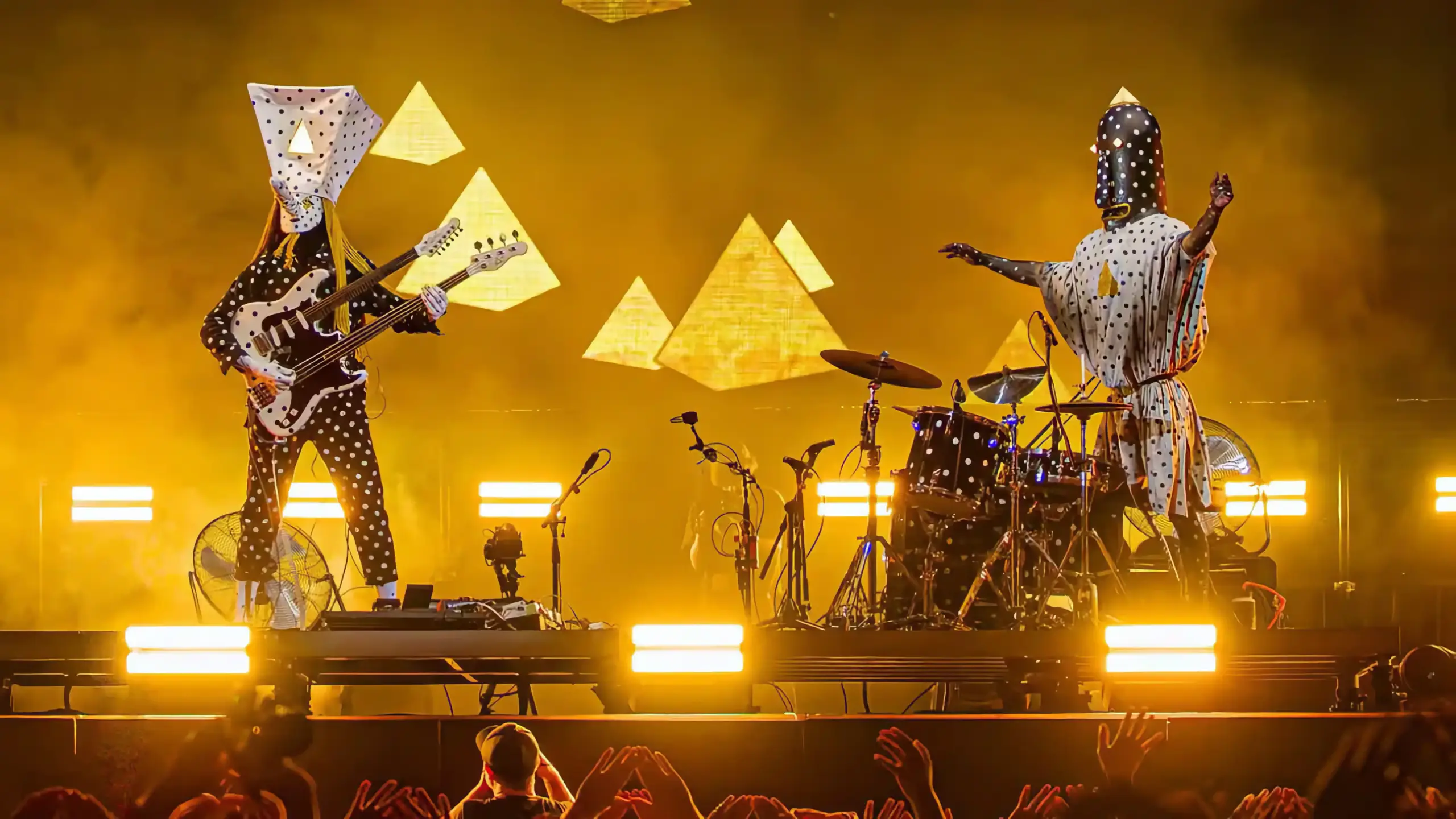 Angine de Poitrine performing on stage in pyramid-shaped papier-mâché head costumes under gold stage lighting.