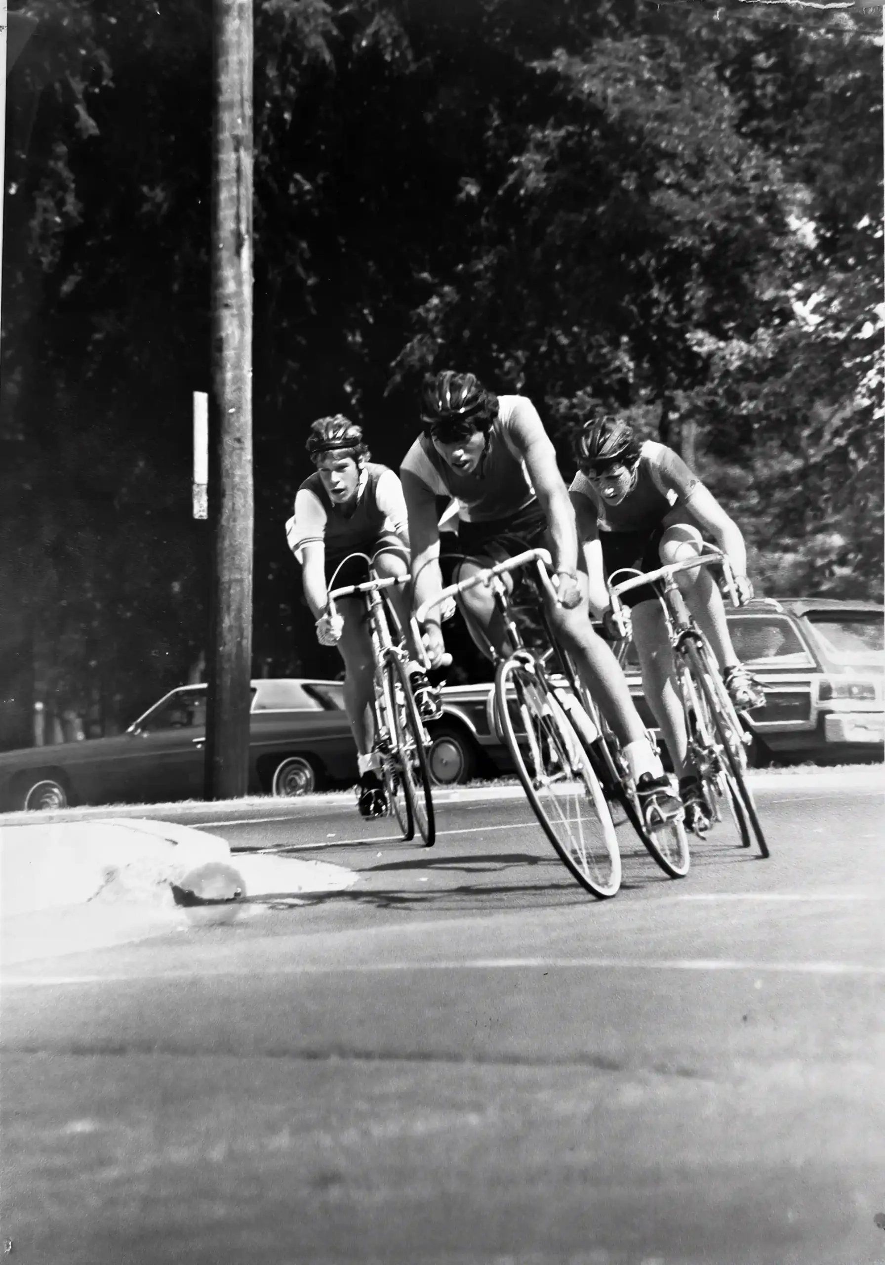 Patrick Oancia racing on a bicycle alongside two other cyclists on a tree-lined road in Halifax, Nova Scotia, 1979.